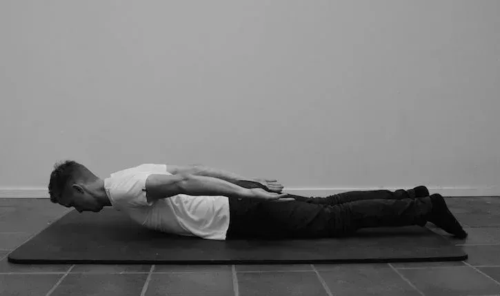 Mann ligger på mage på matte med armene over hodet, og utfører en ryggstrekkøvelse.