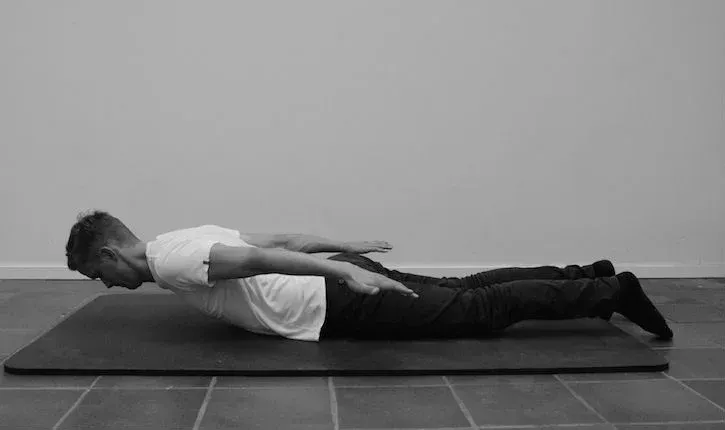 Mann på magen, med utstrakte armer, utfører en ryggøvelse på en matte.