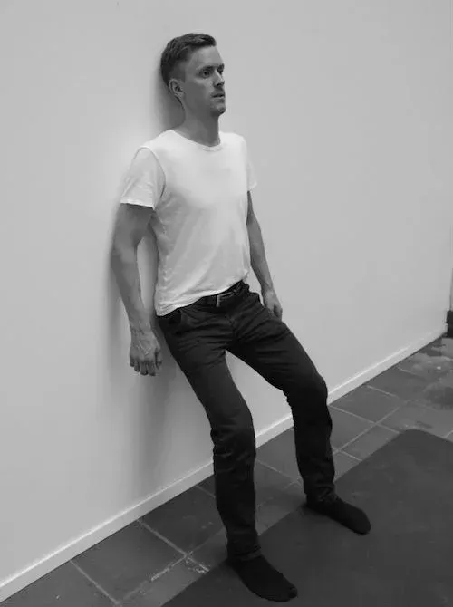 Mann i en vegg sitter med ryggen mot en hvit vegg. Han har på seg en hvit skjorte og mørke bukser, innendørs.