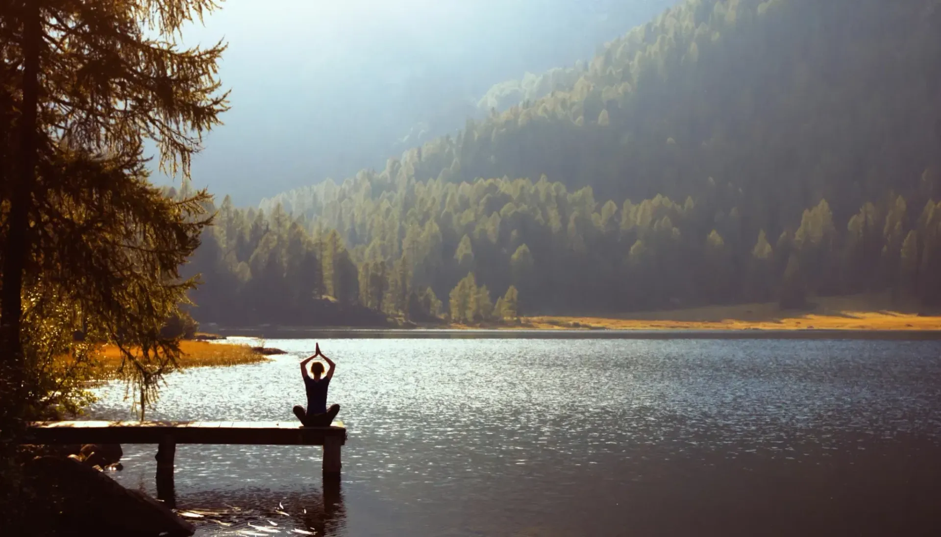 Person som mediterer med armene hevet på en trebrygge med utsikt over en innsjø og fjell i sollys.