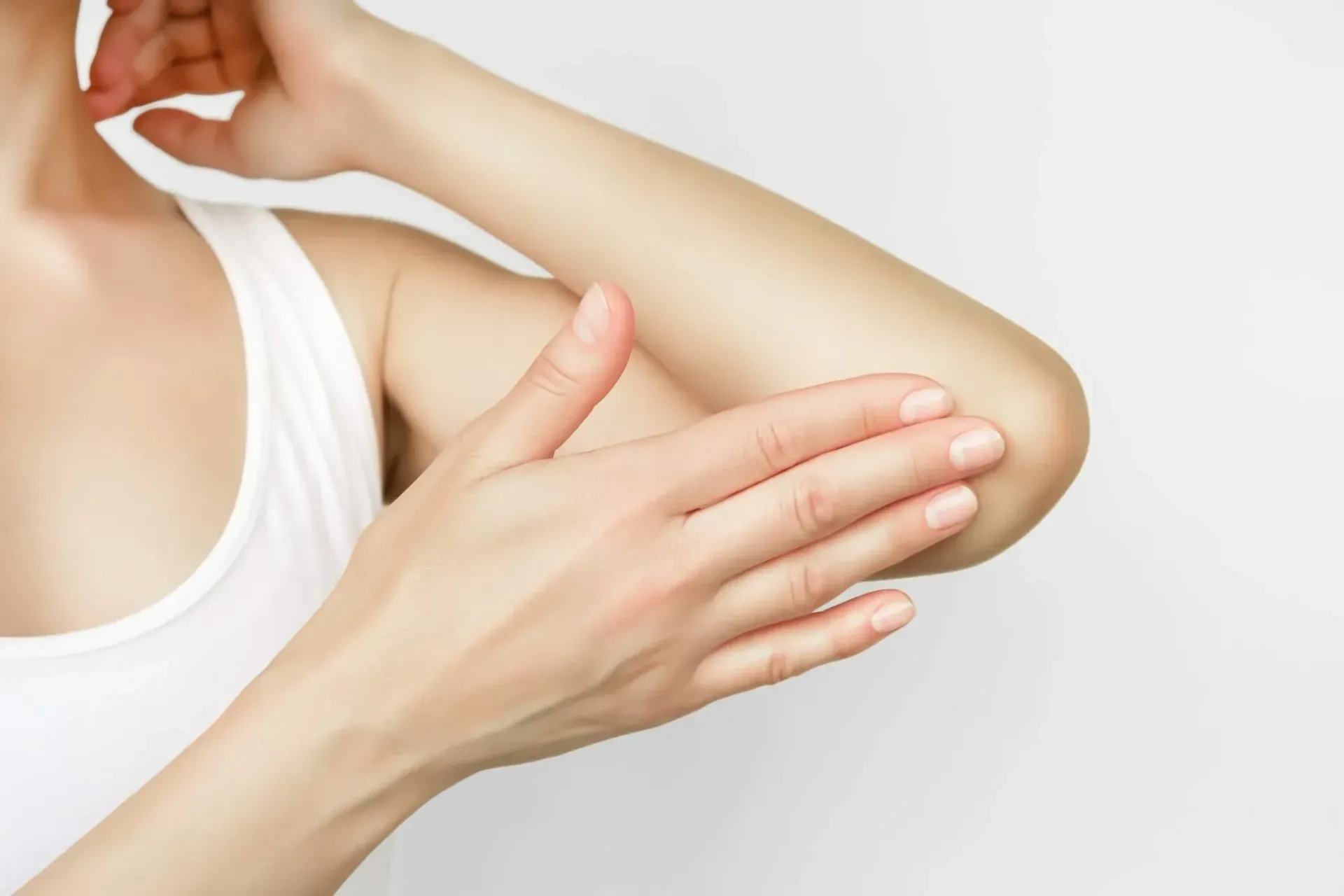 Kvinne som berører albuen sin, iført en hvit tanktopp, mot en hvit bakgrunn.