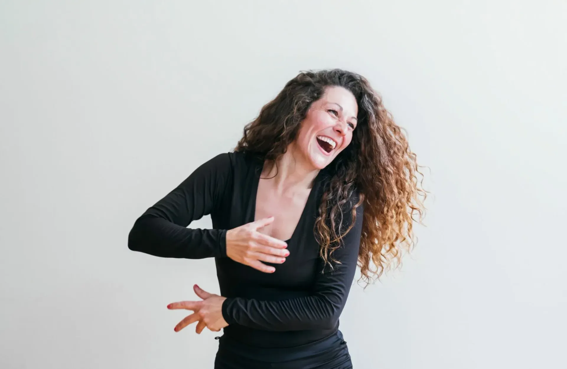 Kvinne som ler, med utstrakte armer, krøllete hår, svart topp, mot en hvit vegg.