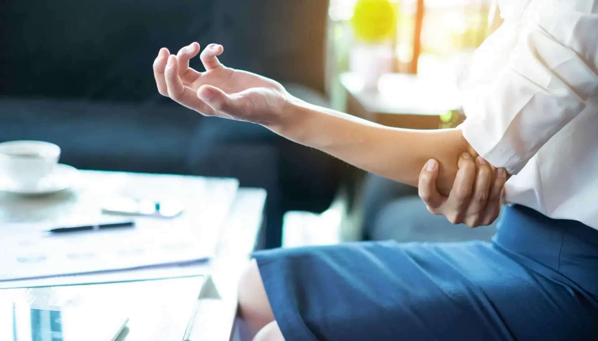 Kvinne som holder en arm, ser ut til å ha smerter i albuen. Sitter nær et bord, i naturlig lys.