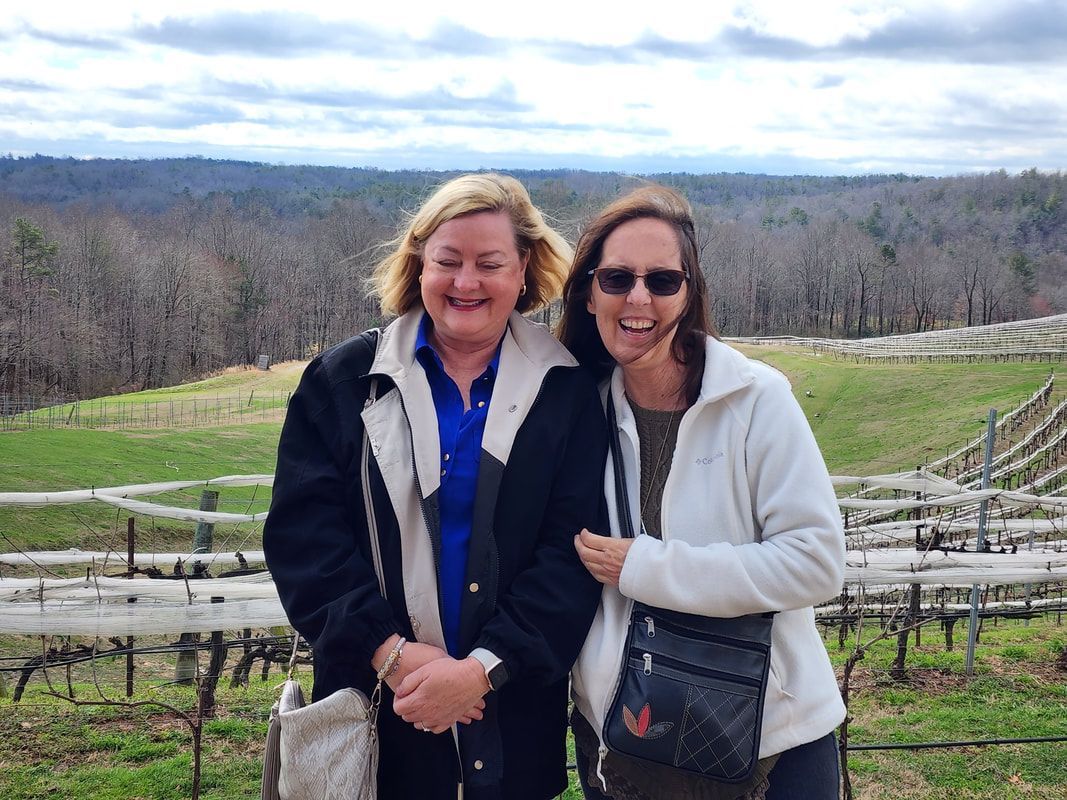Two women are posing for a picture in front of a vineyard.