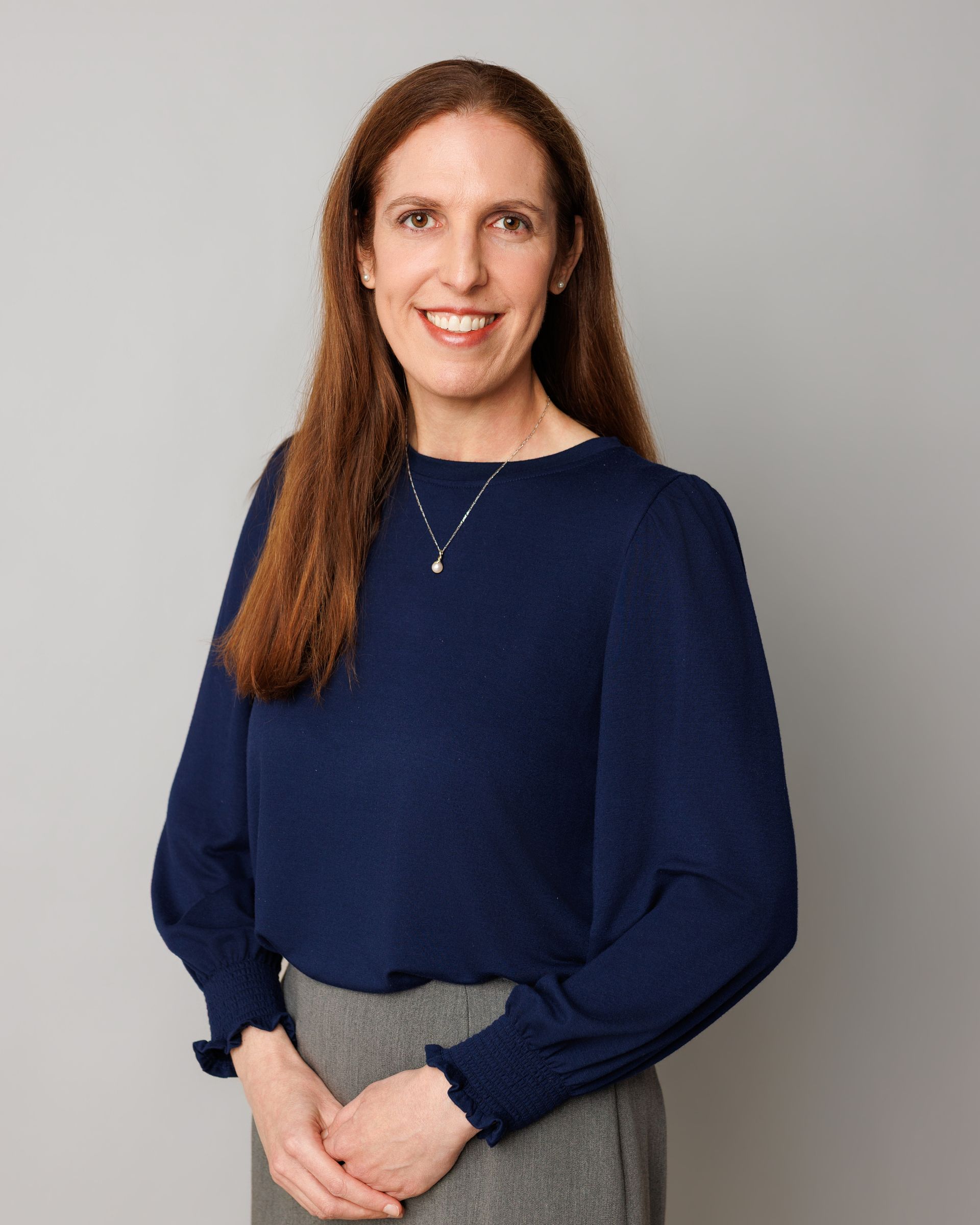 Woman with long brown hair smiles at the camera while wearing a dark blue top, a silver necklace, and small earrings. Outdoor setting with trees.