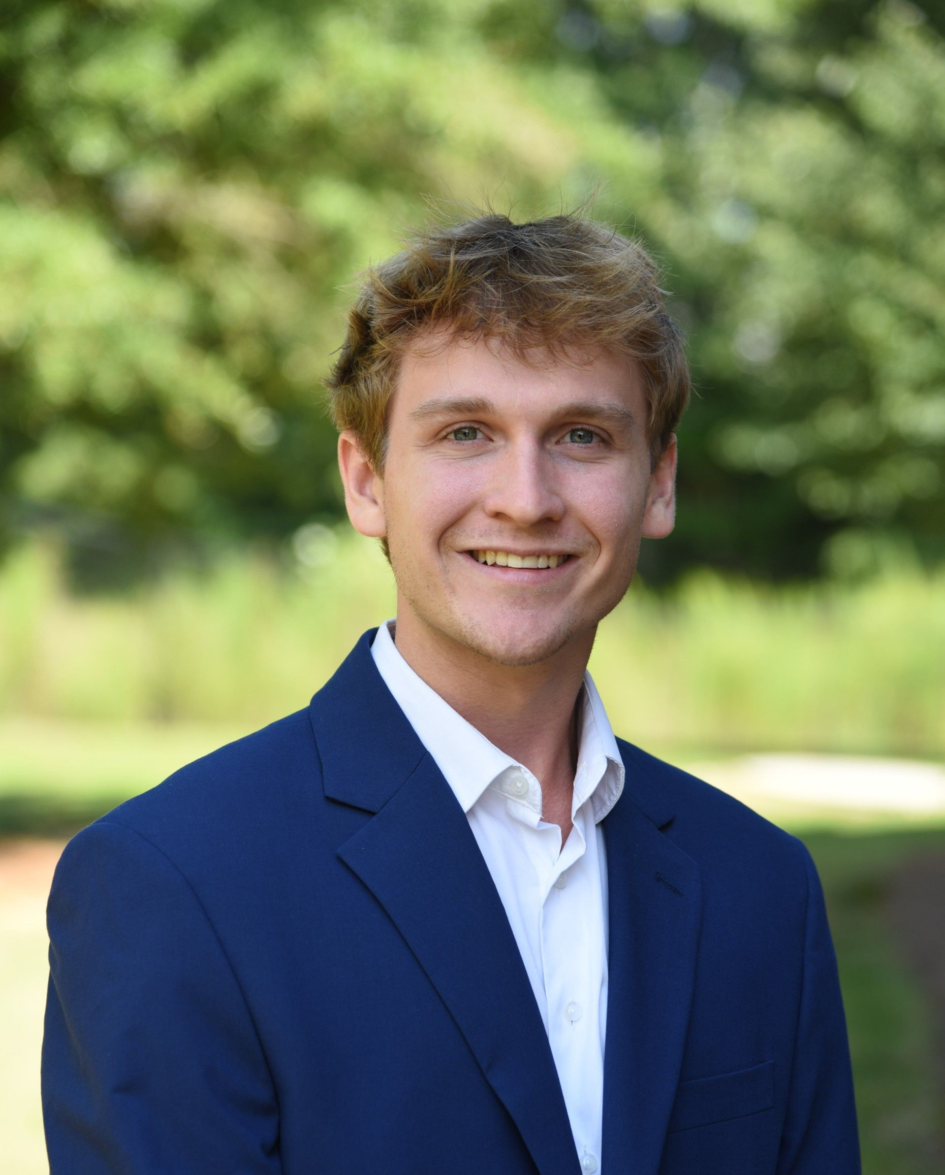 Young man with blond hair in a blue suit and white shirt smiles outdoors with a blurred green background.