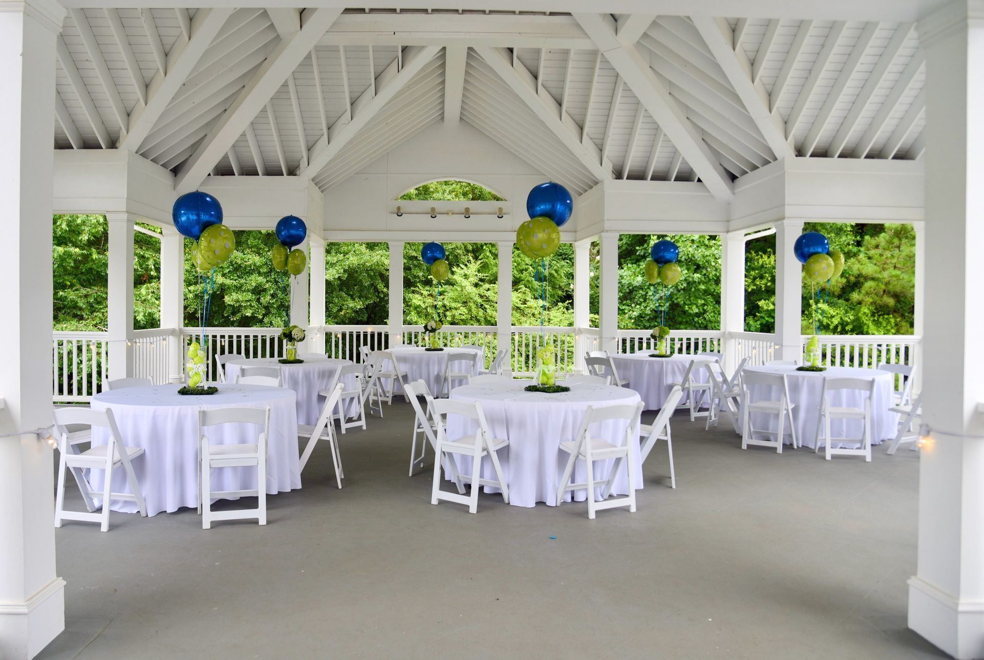 White gazebo set for an event with round tables covered in white tablecloths, chairs, and blue and green balloons.