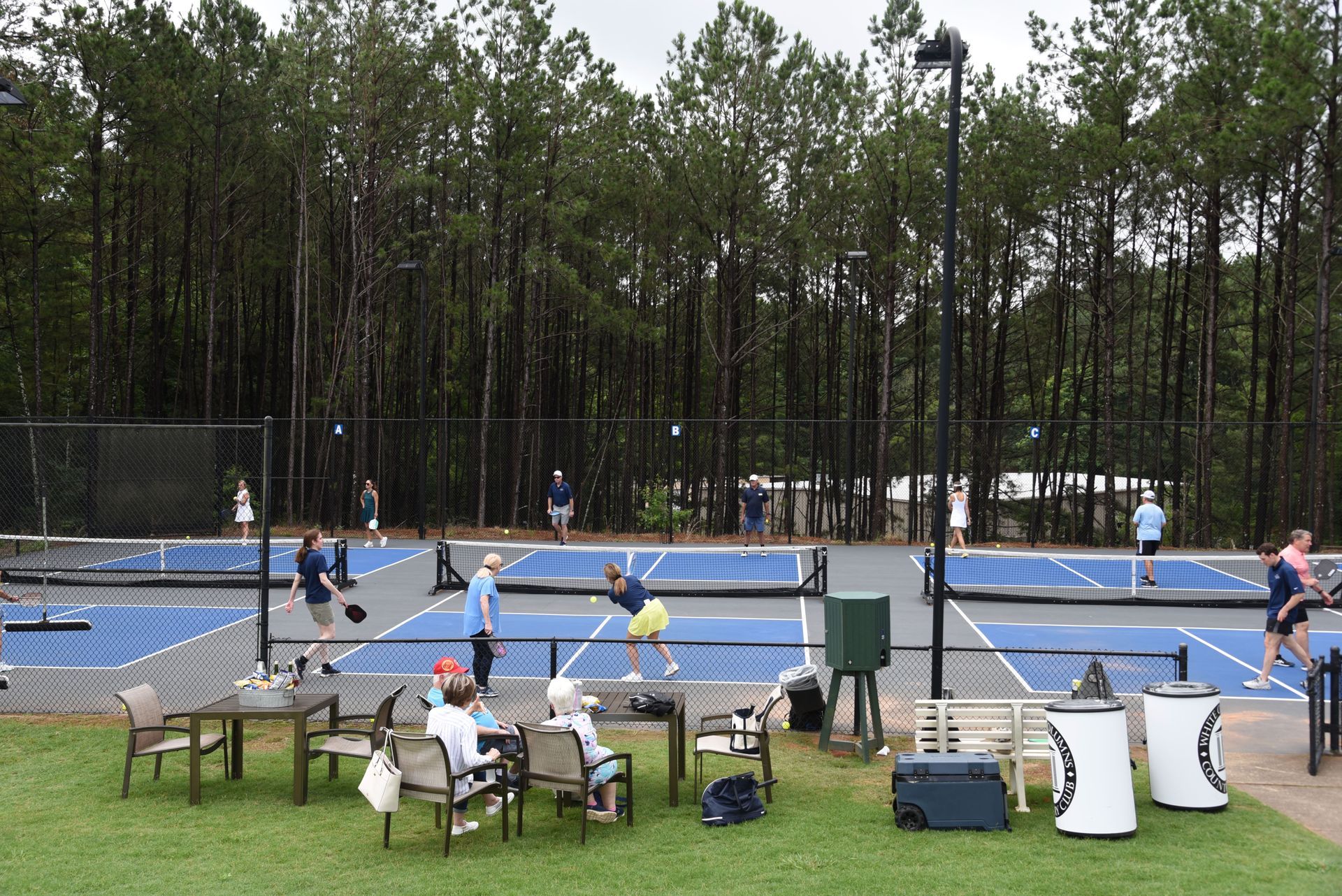 Pickleball players on a blue court, watched by people at tables. Trees in the background.