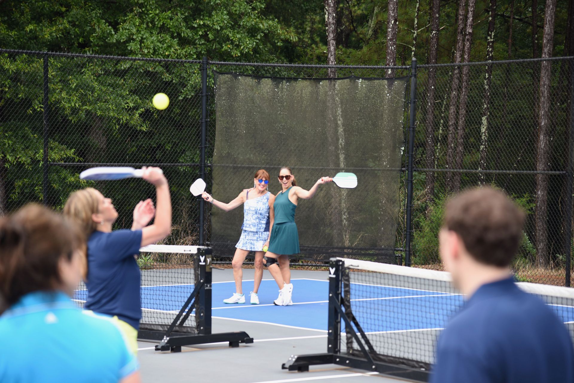 People playing pickleball on a blue court. Two women in the middle hold paddles up, while others play and watch in a court near a fence and trees.