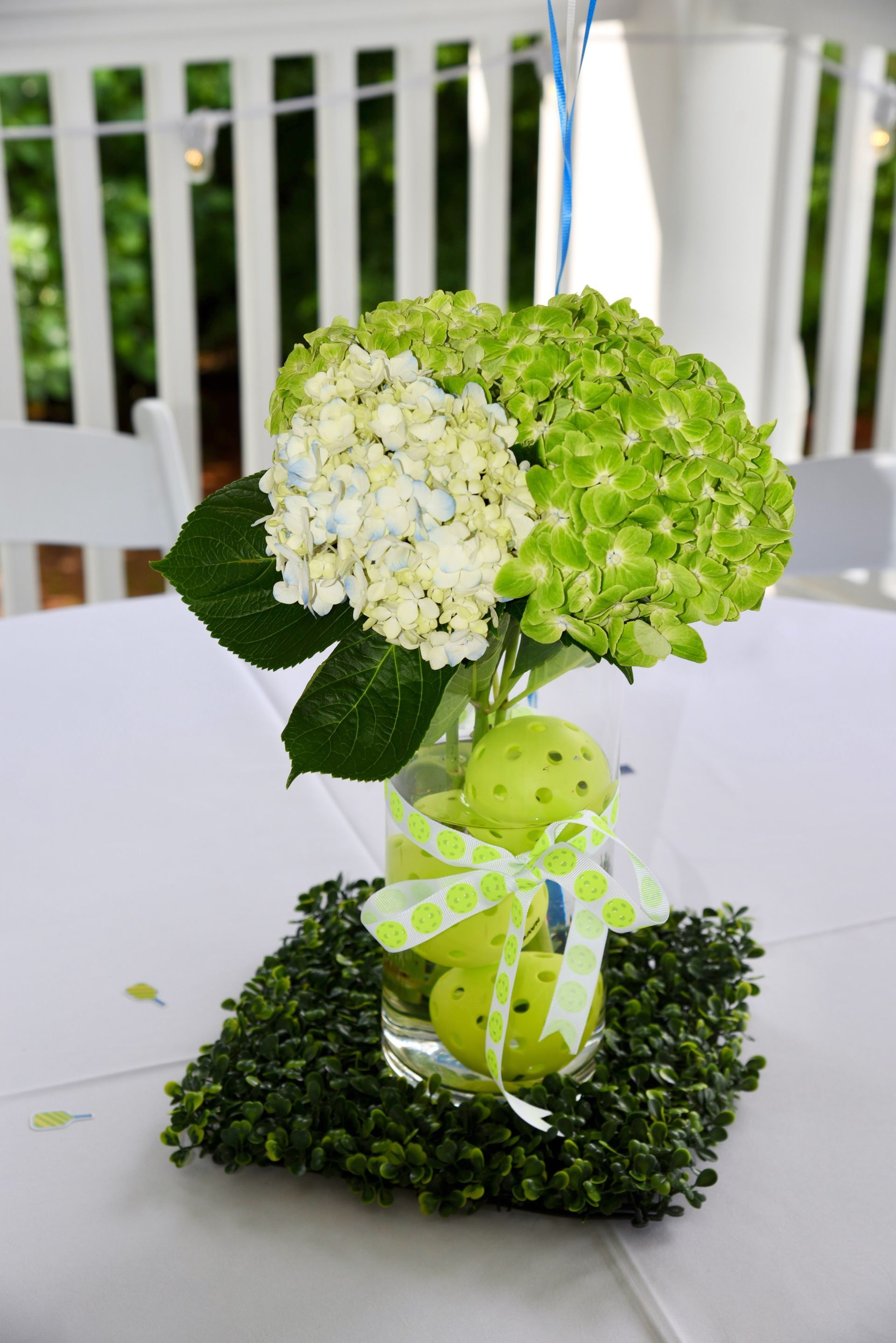 Floral centerpiece with green and white hydrangeas in a clear vase containing green apples tied with a light green polka dot bow, resting on a square bed of greenery, on a white tablecloth.