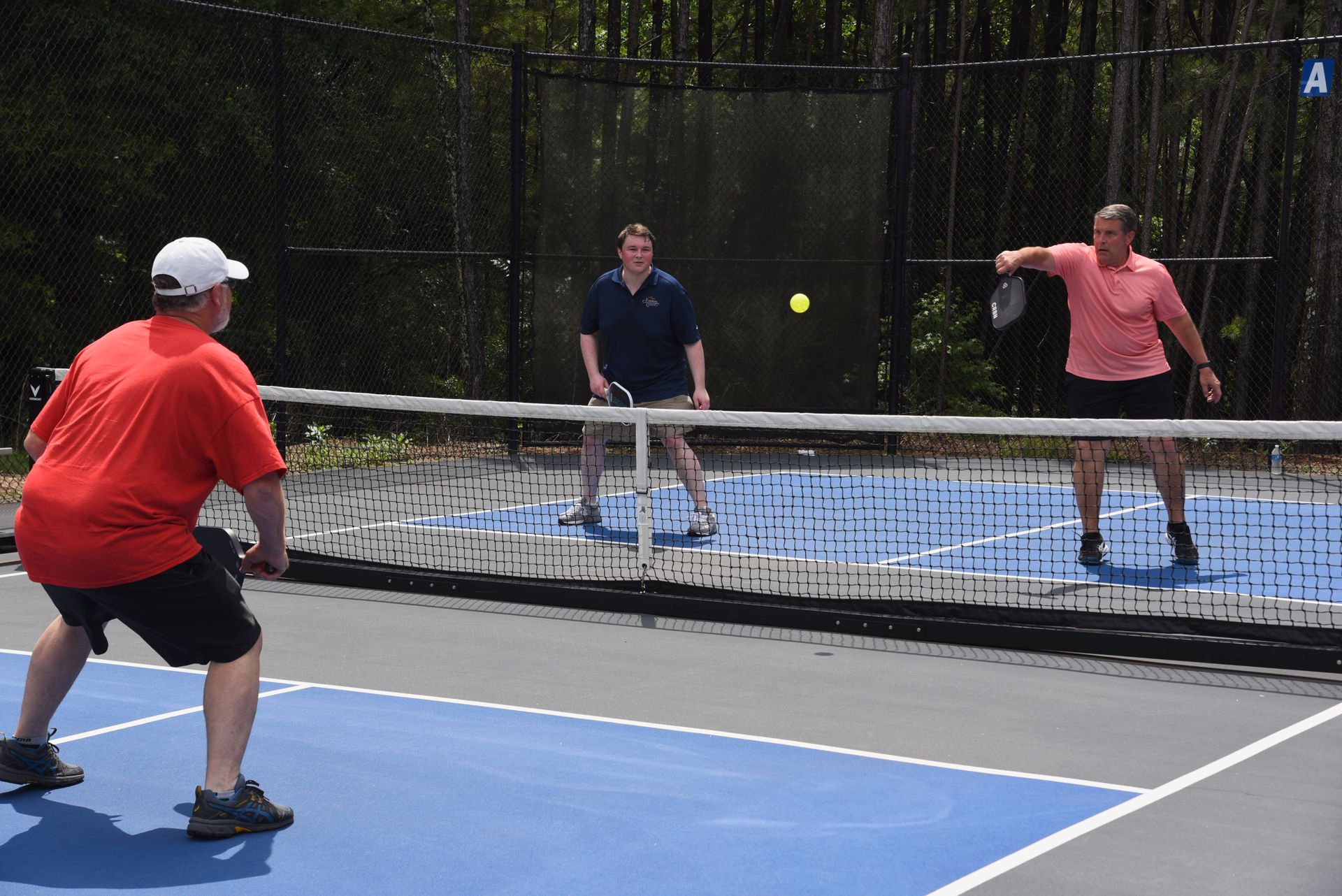 Four people playing pickleball on a blue court. One player in a red shirt is ready to hit the ball, while others watch and prepare to play.