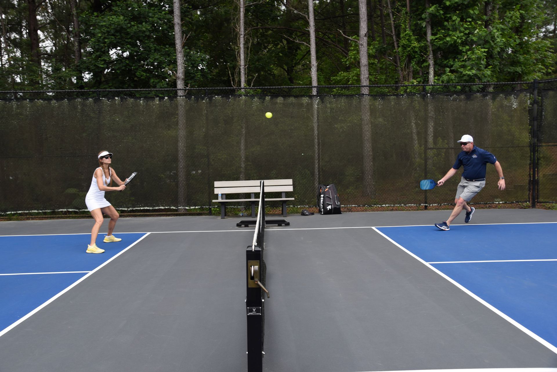 Two people playing pickleball on a blue court. A woman in white is ready to hit the ball, while a man moves forward to return it.