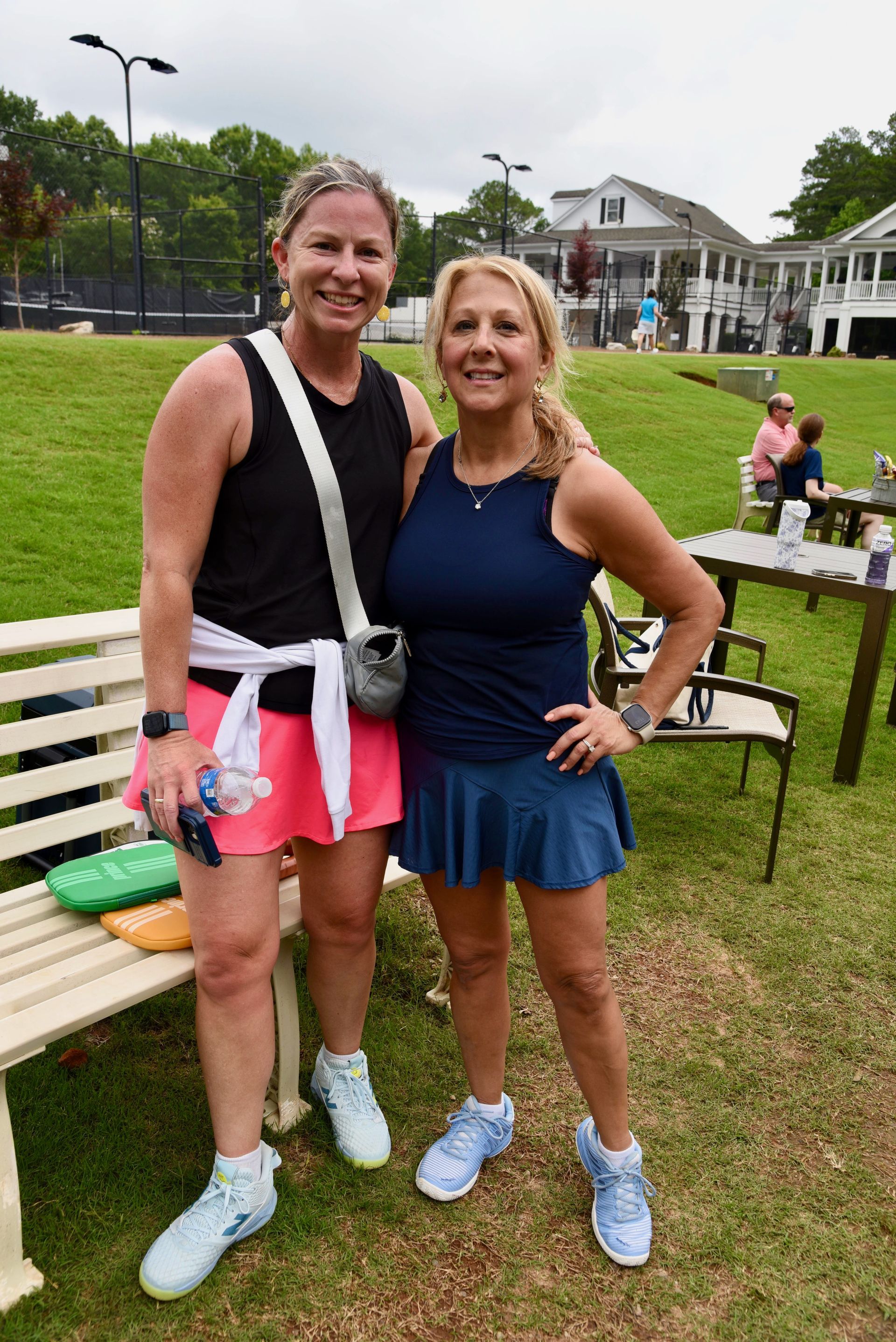 Two women stand together on a grass court, wearing athletic skirts and tops. One has a white jacket tied around her waist.