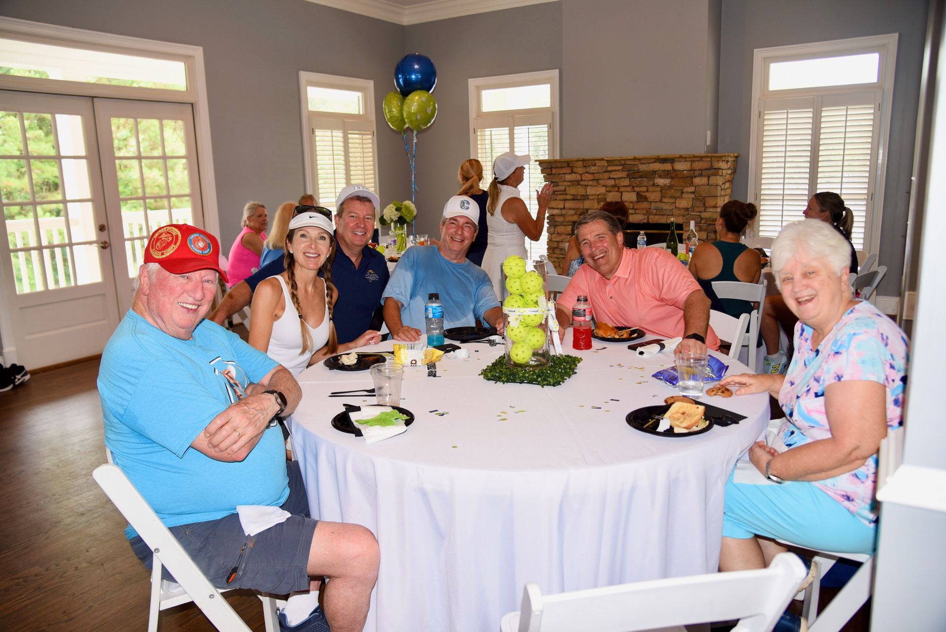 Group of people seated around a round table in a bright room; they smile and eat, with balloons visible.