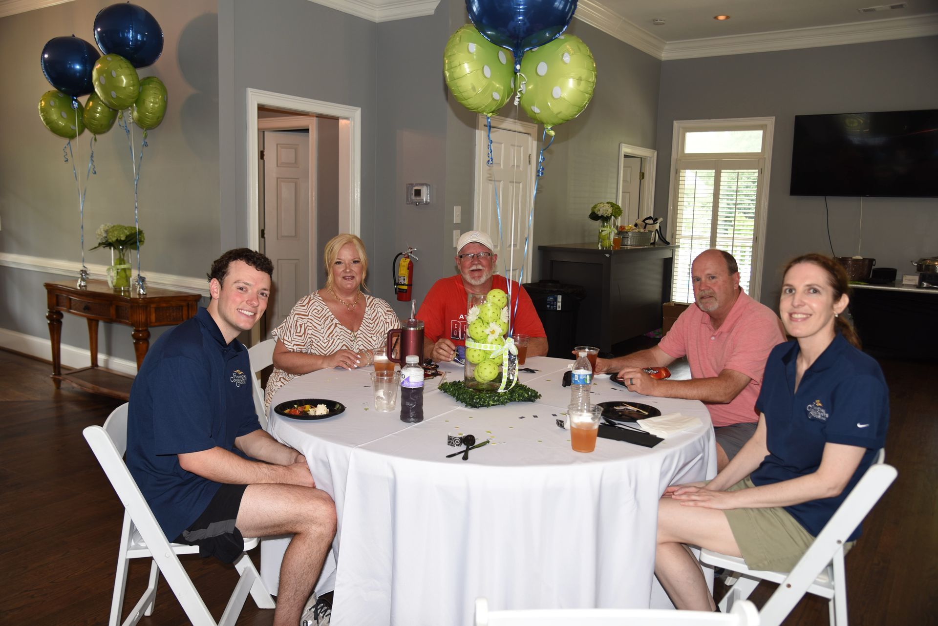 Five people seated around a round table decorated with balloons. They appear to be at a celebratory event.