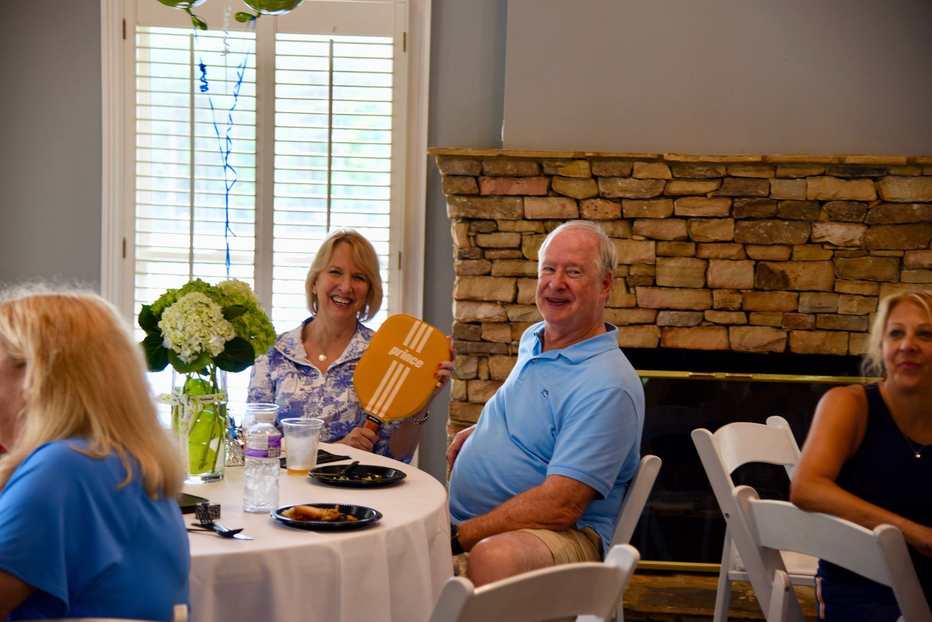 A man and woman smile at a round table. The man holds an orange fan near a stone fireplace.