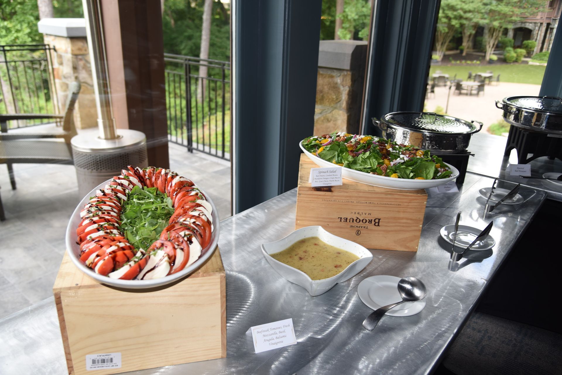 A buffet table with salads and dressings, set outdoors near a patio. Tomato and mozzarella salad is on the left, with a greens salad behind.