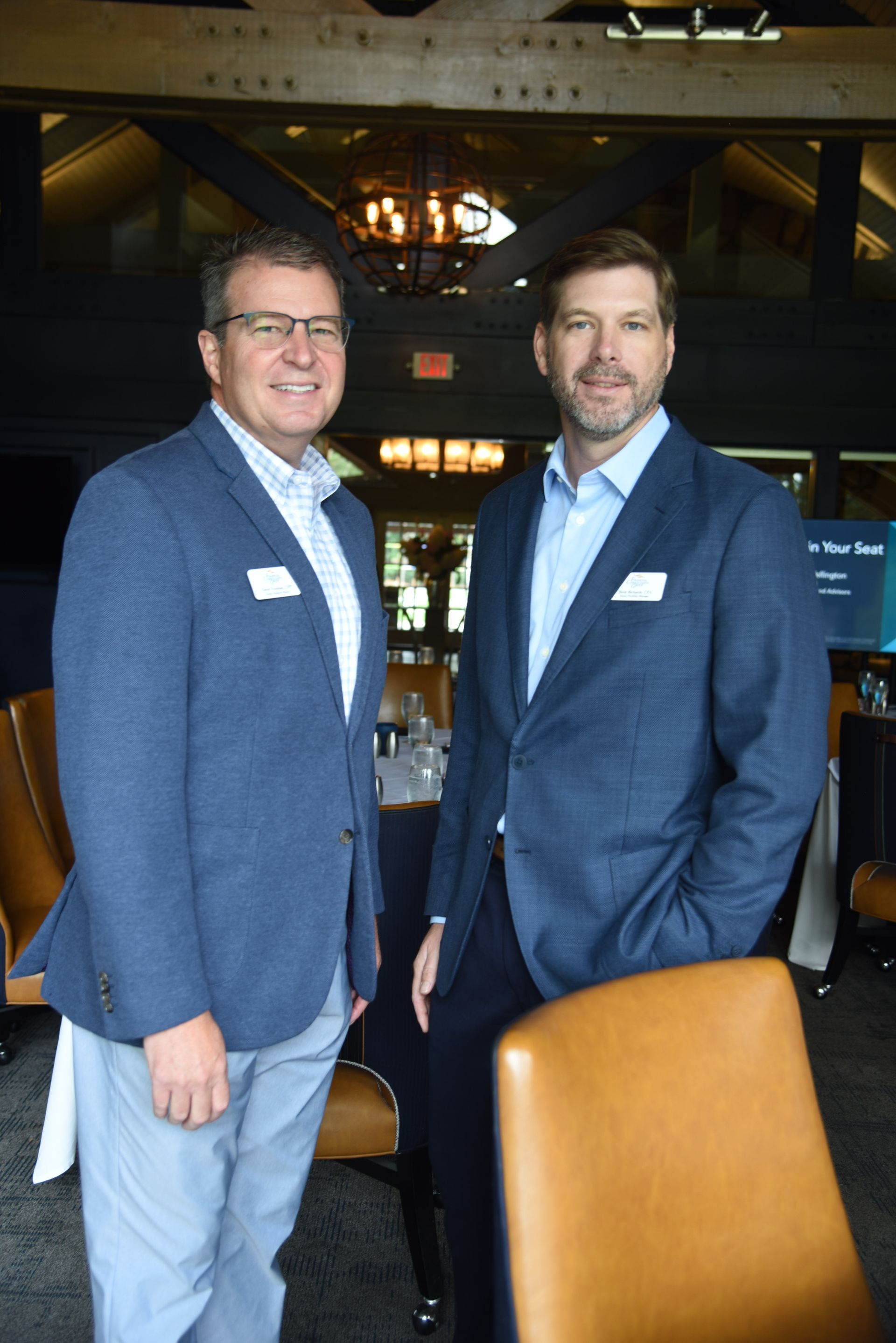 Two men in blue blazers and dress shirts stand together. They are in an upscale setting with leather chairs and a dark background.