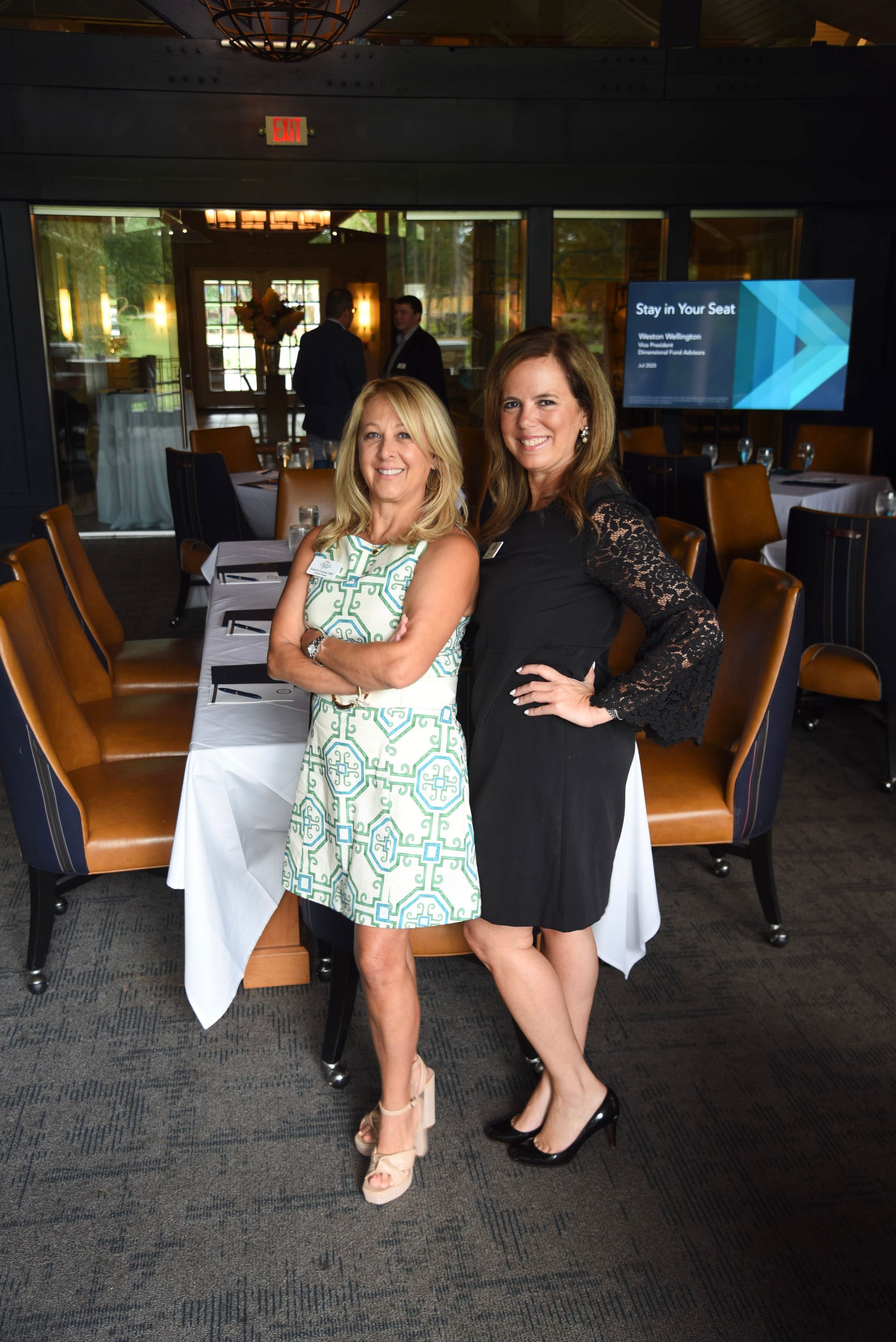 Two women smiling, posing together in a restaurant. One wears a floral dress and wedges, the other a black dress and heels.
