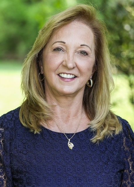 Woman with blonde hair smiles outdoors; she wears a blue top and necklace.