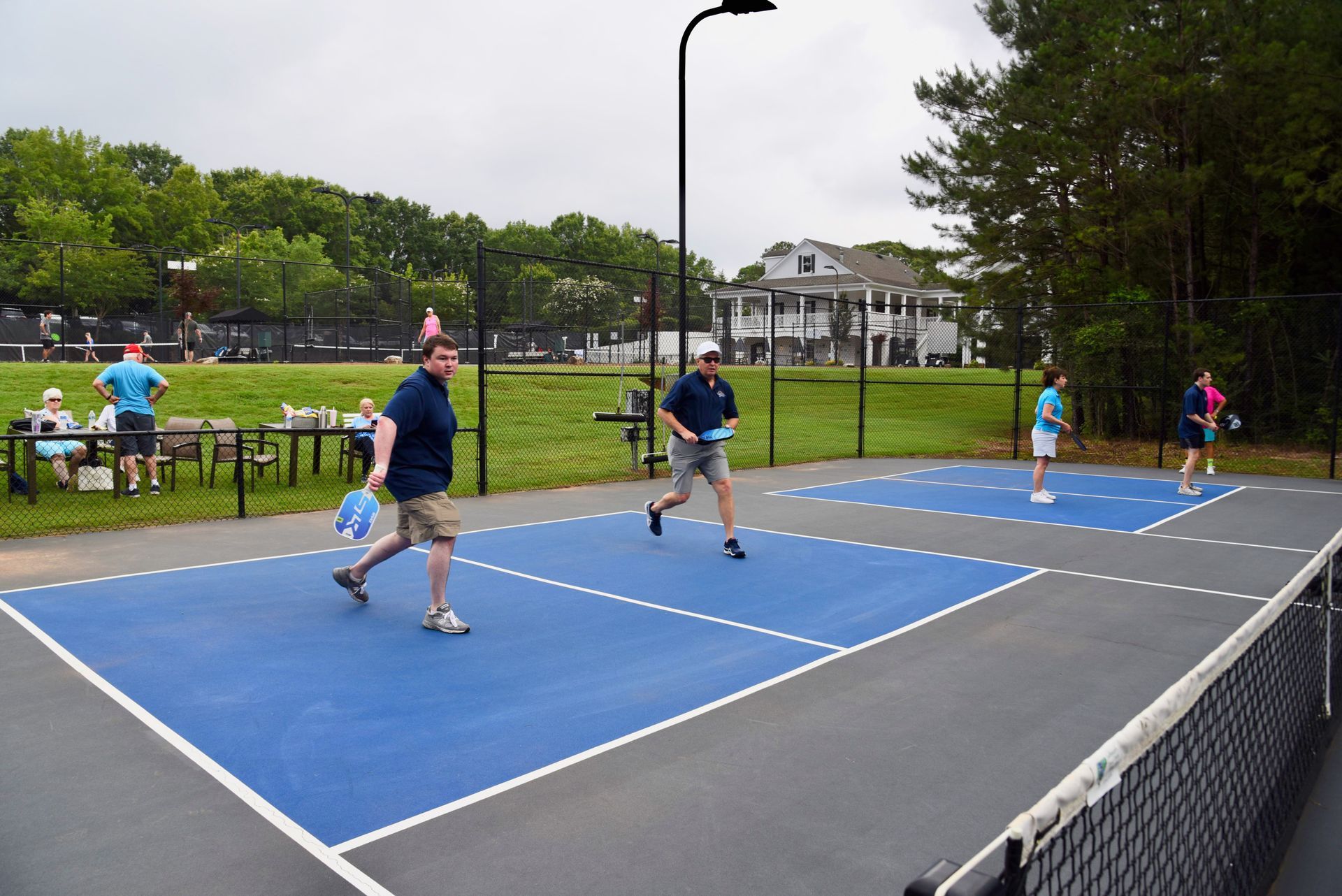 People playing pickleball on an outdoor blue court with a net, under a cloudy sky. Other players watch nearby.