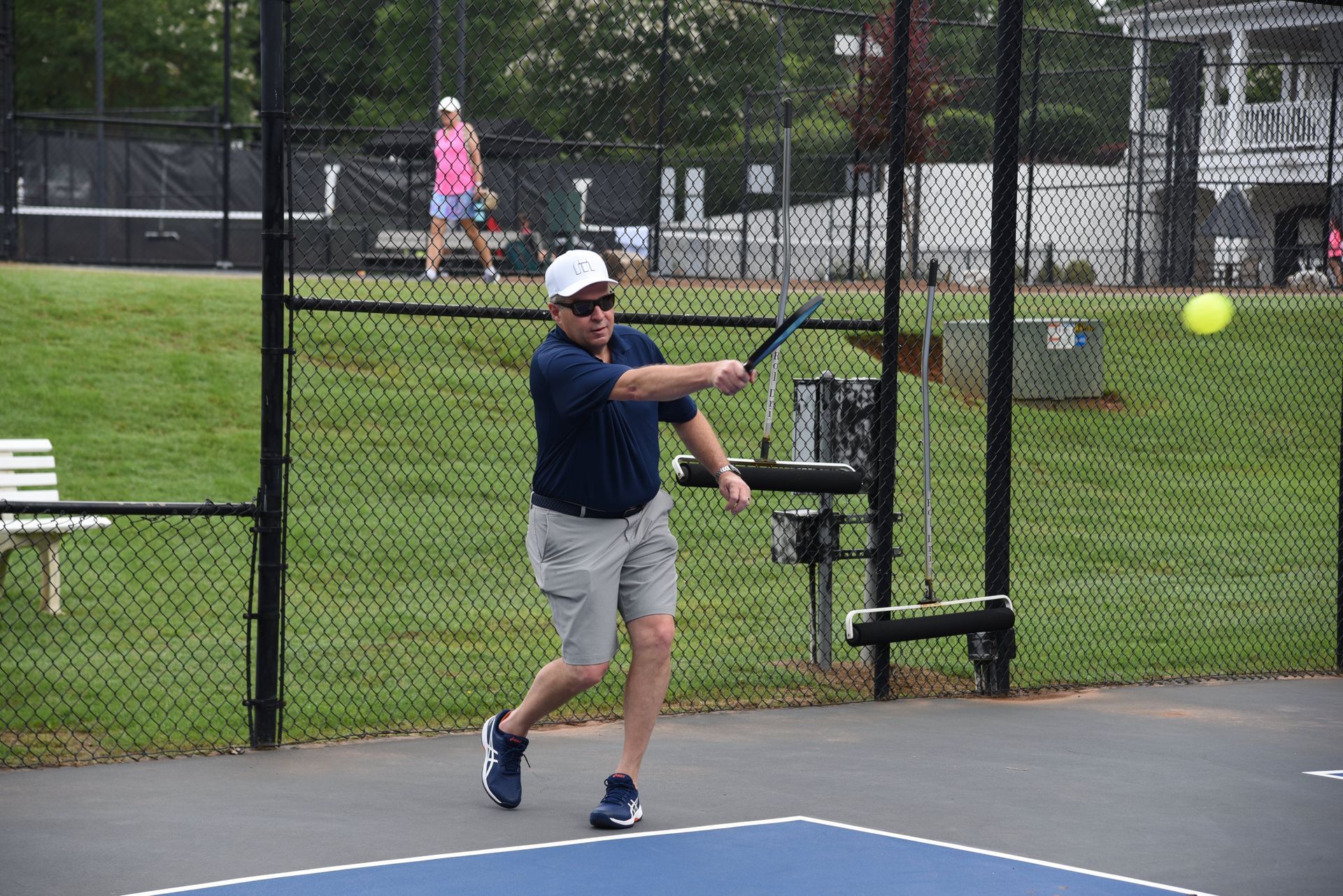 A man in a navy shirt and grey shorts playing pickleball on an outdoor court, swinging at the ball. Another player stands in the background.