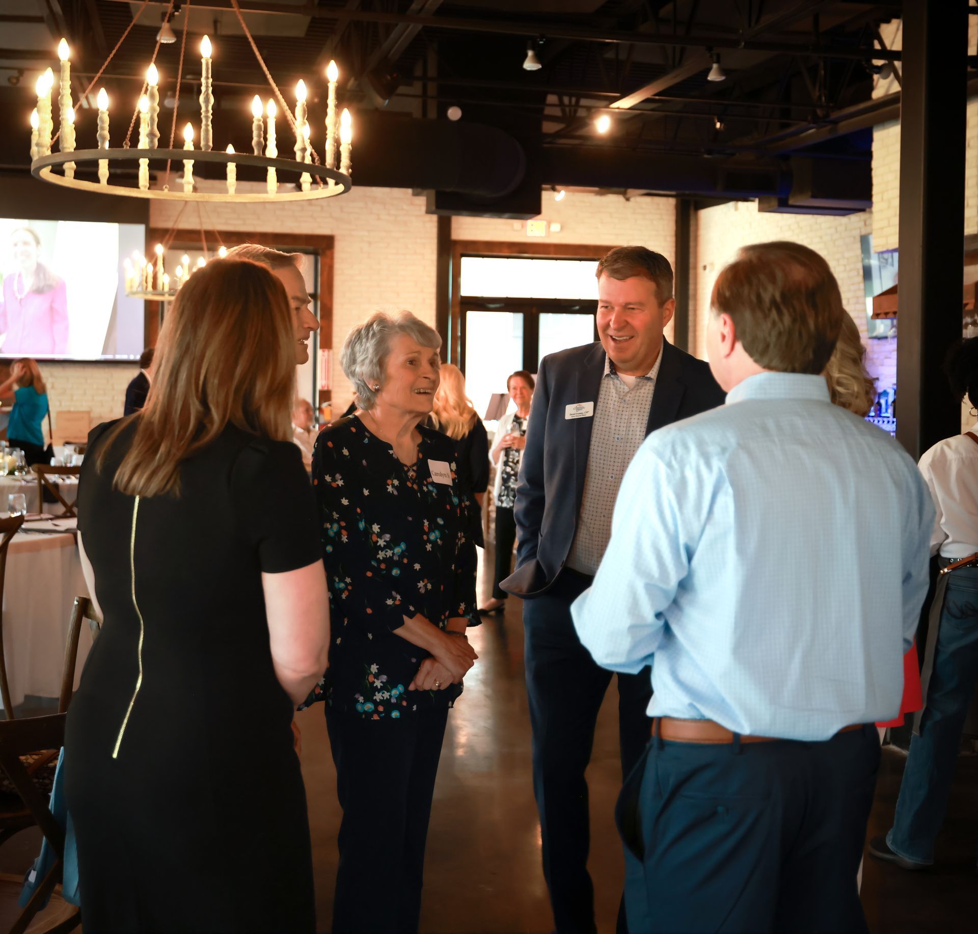 Group of people in a well-lit room socializing. Woman in black dress, man in blue shirt, and others are talking.