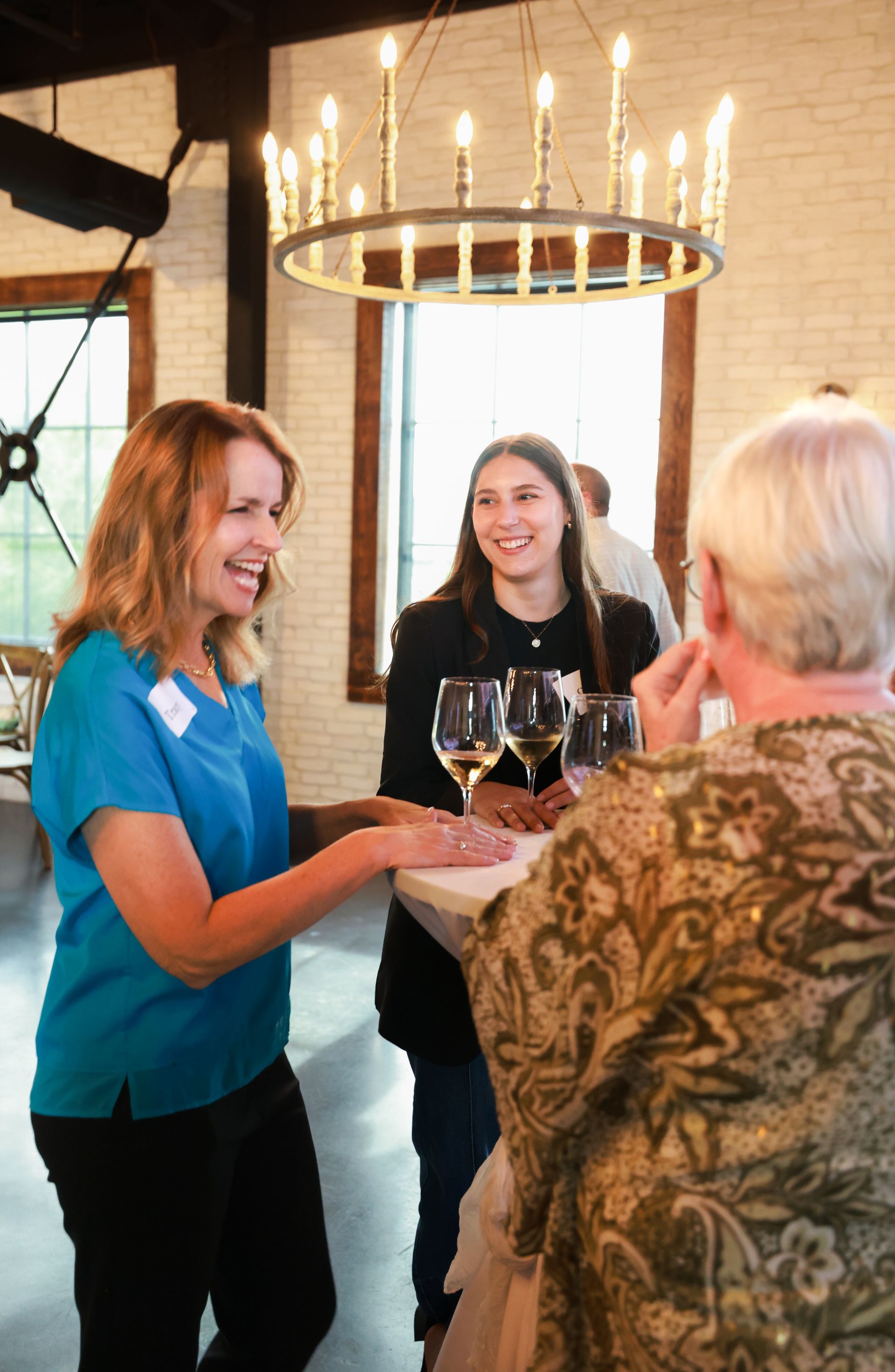 Three women smiling and talking around a small table, holding glasses of wine, under a chandelier in a bright room.