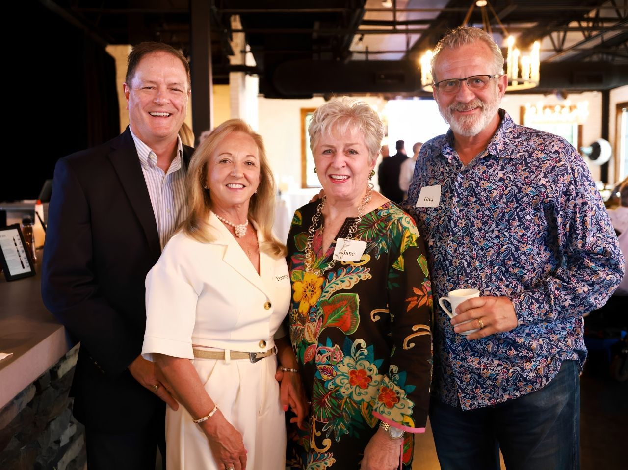 Four people smiling at a gathering: a man in a suit, two women in colorful outfits, and a man holding a mug wearing a patterned shirt.