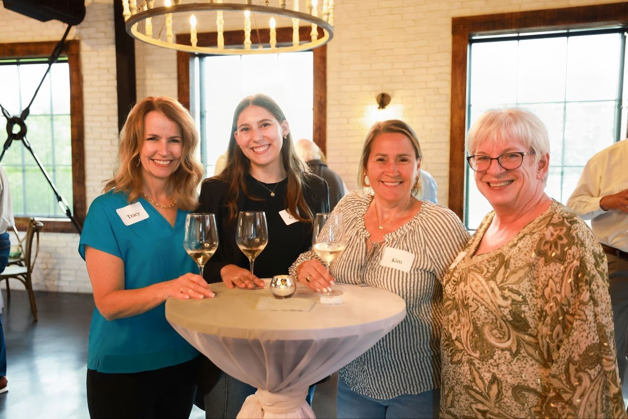Four women smiling and holding glasses of wine at an event, gathered around a small table indoors.