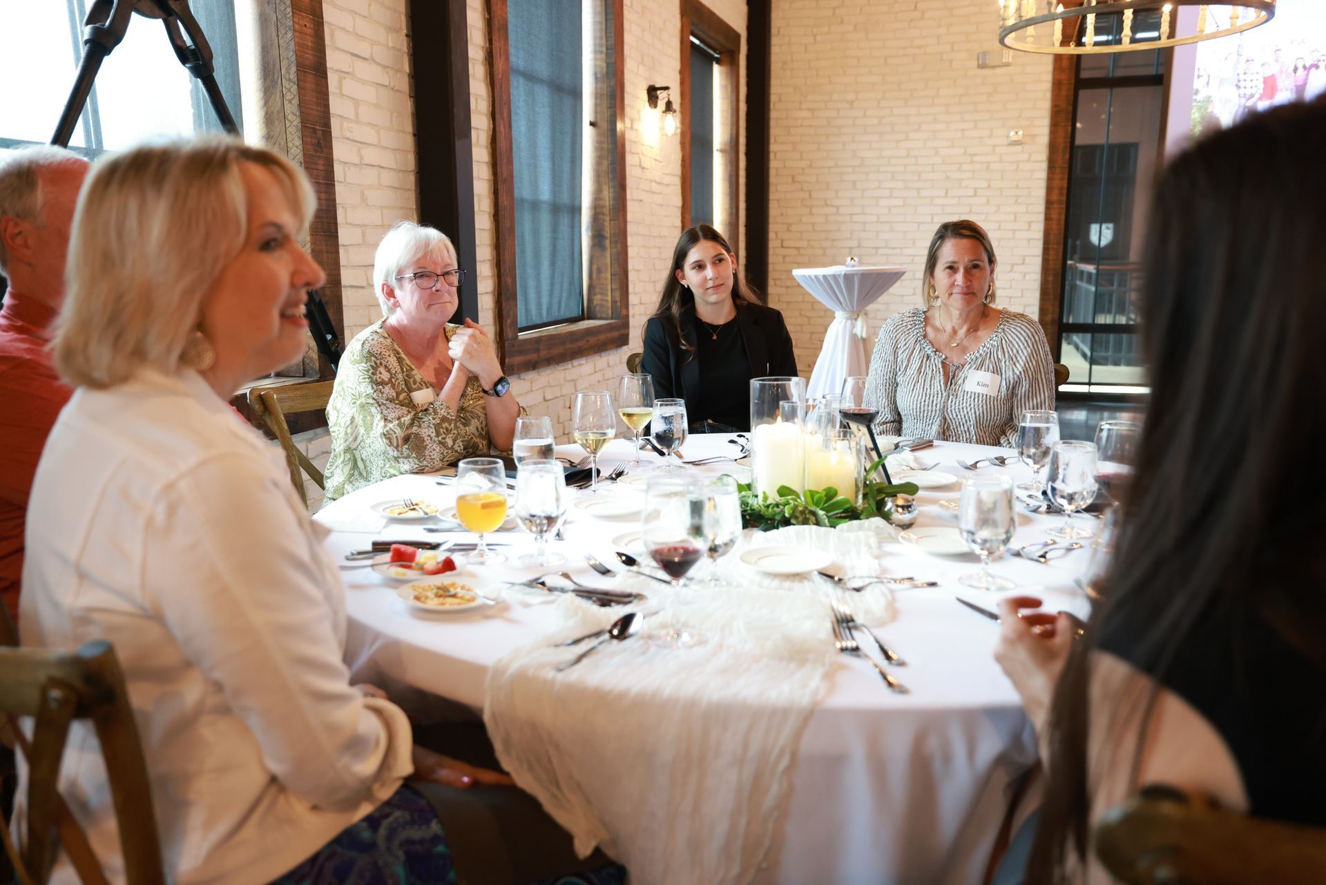 A group of people seated around a white table, eating and conversing. The room has a brick wall and windows.