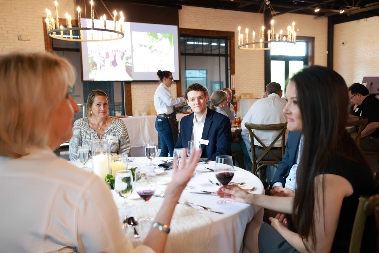 People seated at a round table in a restaurant. Two women are in conversation, while others dine in a bright dining room.