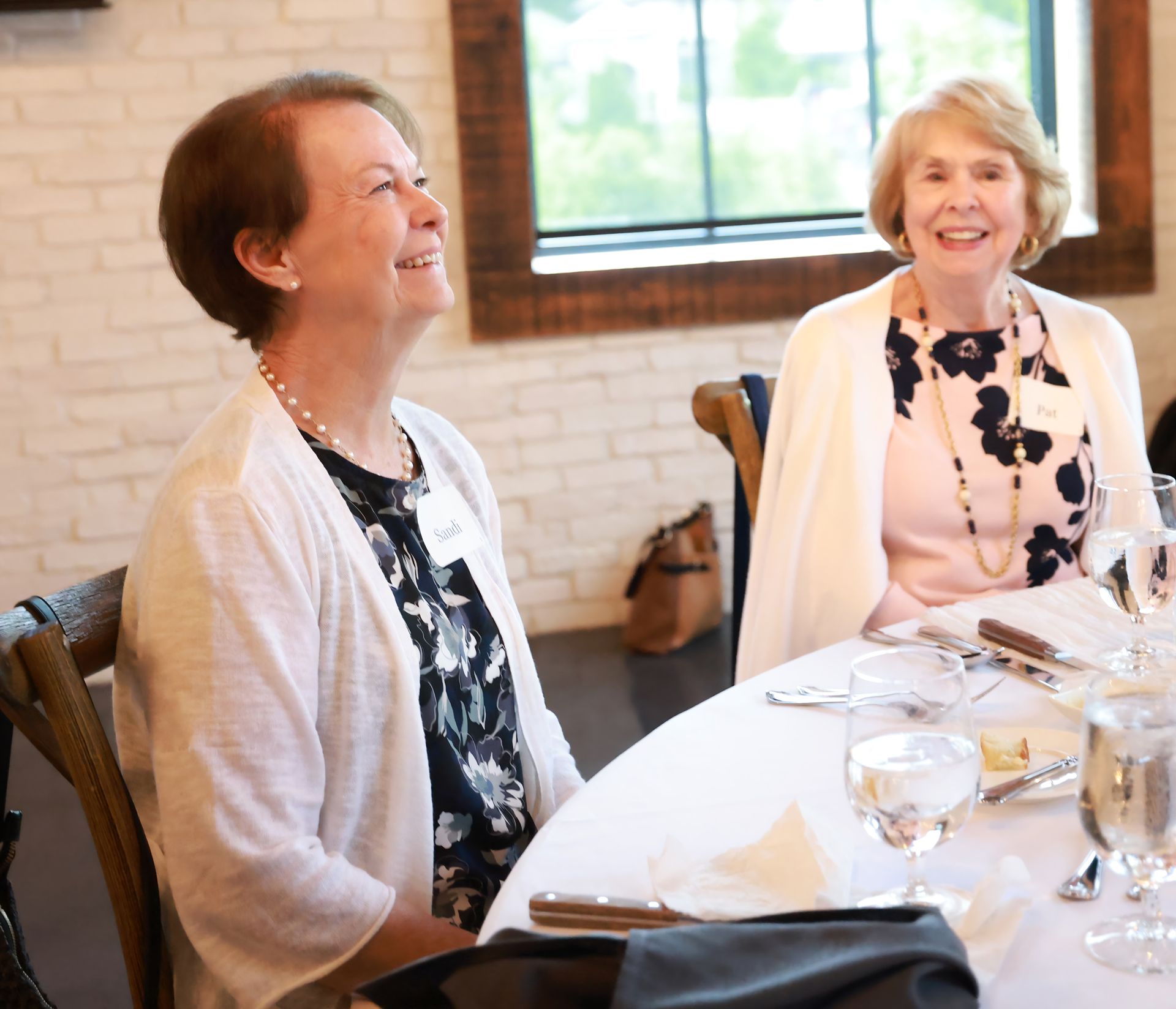 Two smiling women seated at a table in a light-filled room. One wears a floral print and white cardigan, the other a black and white floral top.