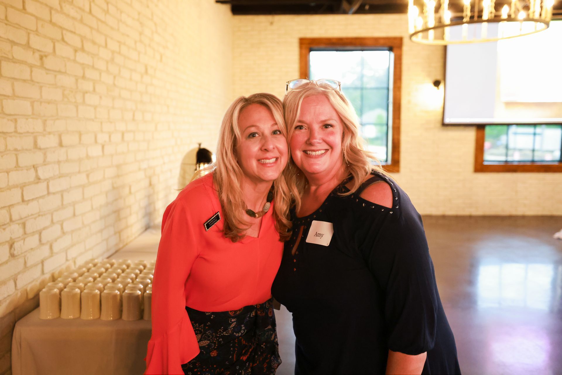 Two smiling women pose for a photo in a room with brick walls. One wears a red top, the other a black top.
