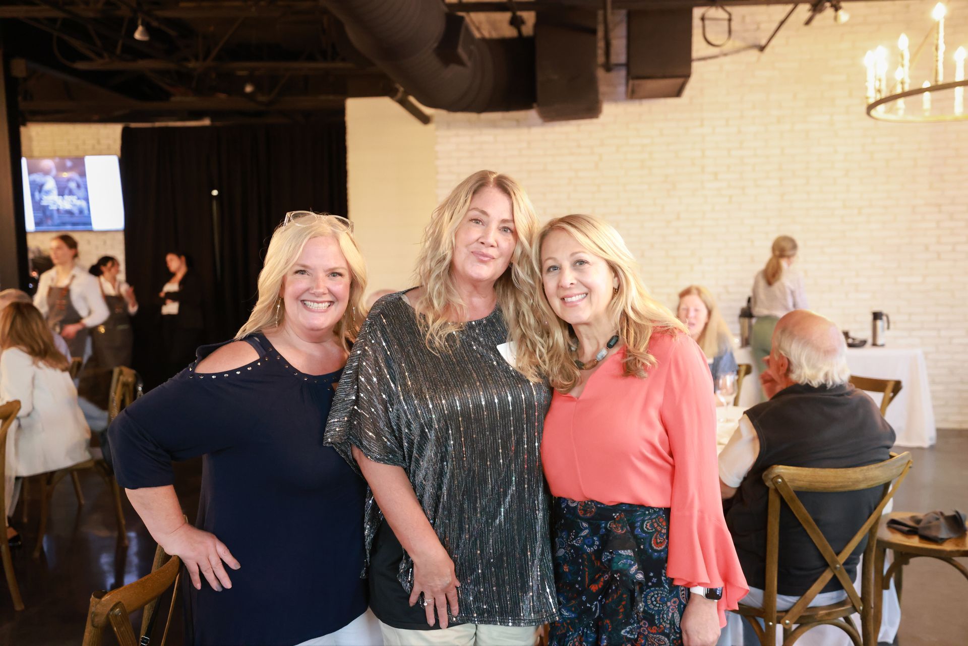 Three smiling women pose for a photo at an event, inside a bright venue with tables and other people.