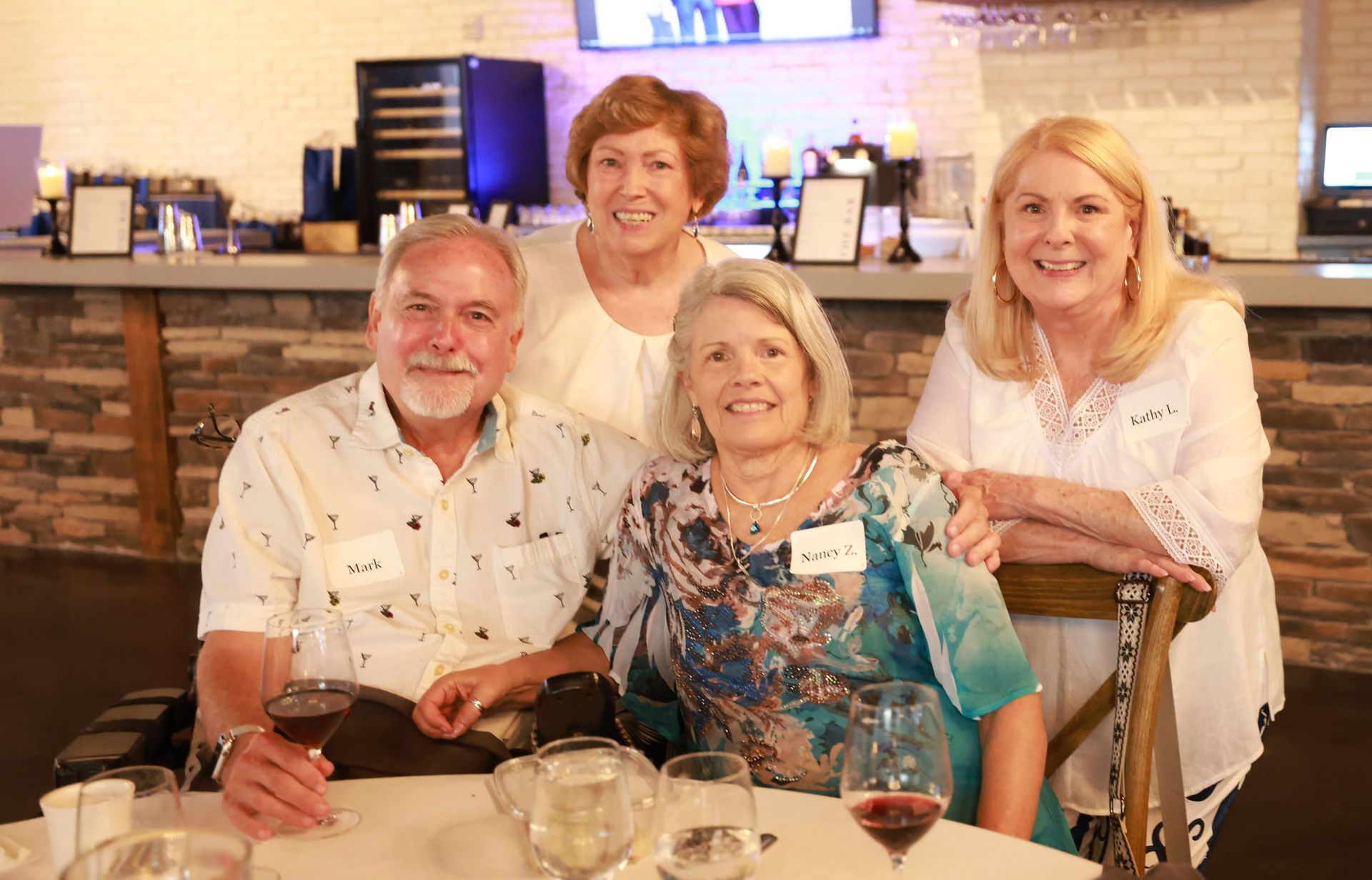 Four smiling adults at a restaurant table. Two seated and two standing, posing with drinks. Bright, airy setting.