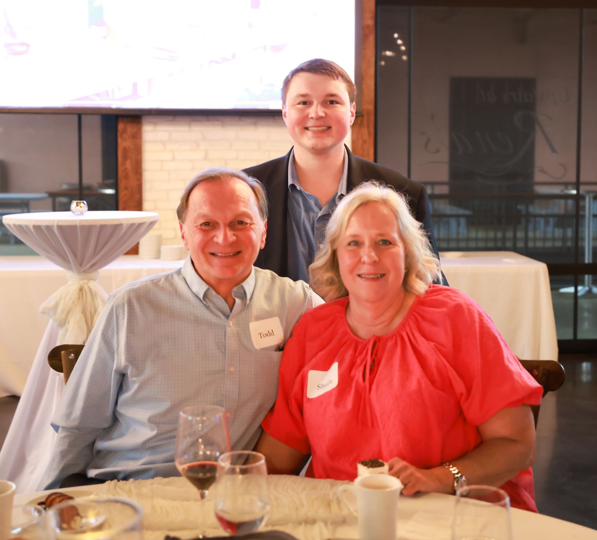 Three people at a table: an older man and woman smiling, a young man standing behind them. They're in a room with tables and a projection screen.