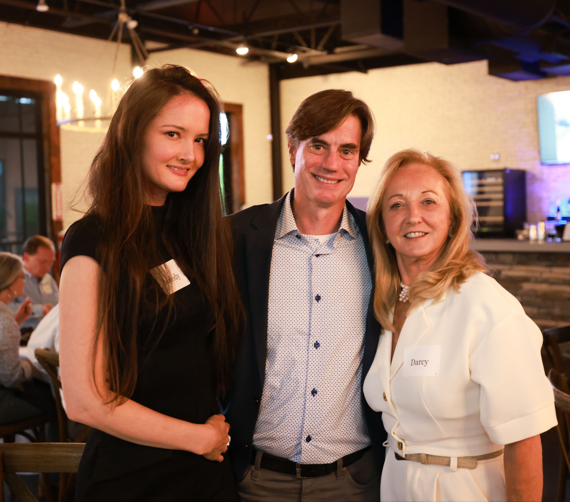 Three people smiling at a casual event. A young woman in black, a man in a blazer, and an older woman in white clothing.
