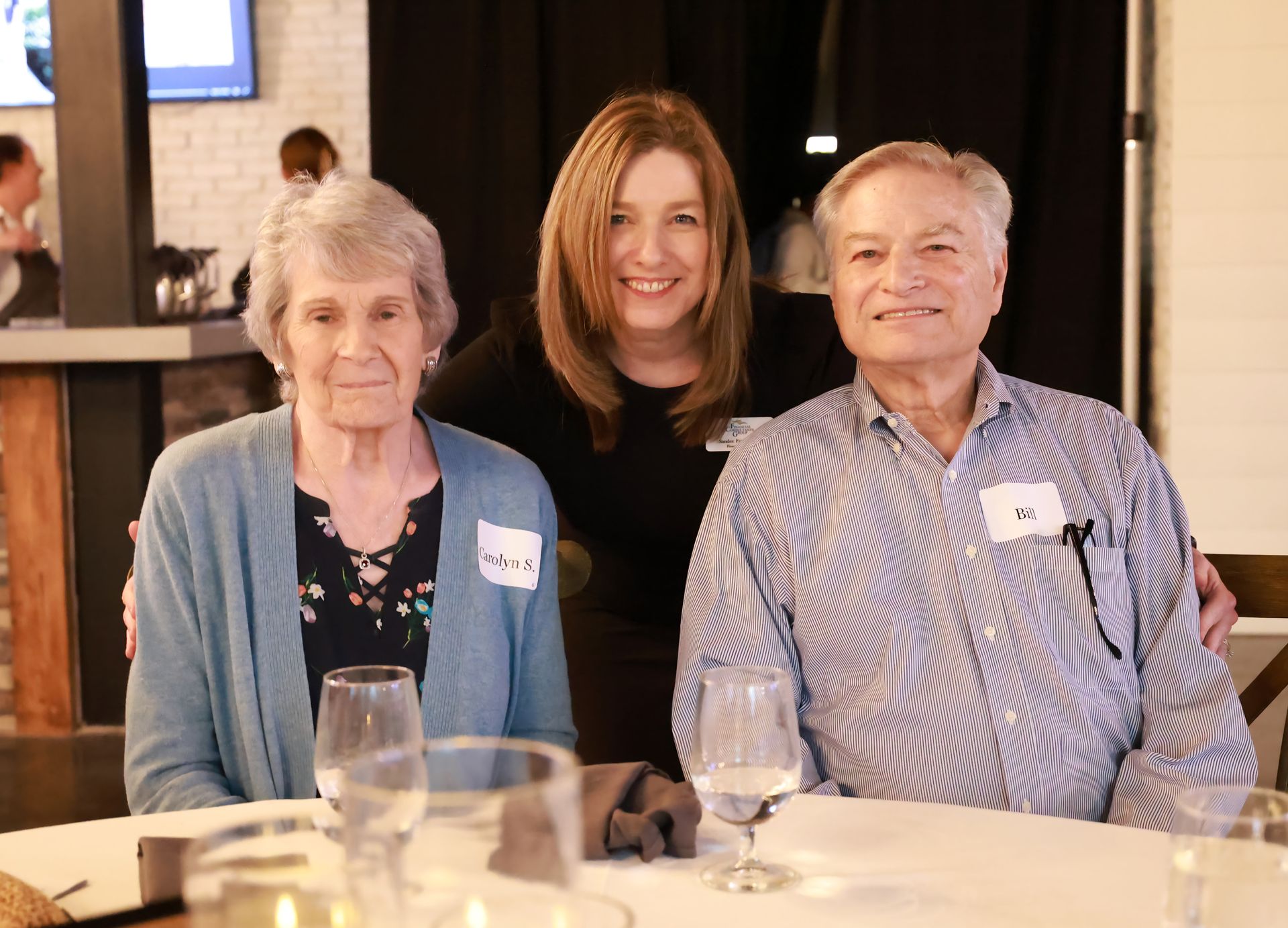 A woman smiles with an elderly couple at a restaurant table. The woman in the middle has brown hair; the couple sits on either side of her.