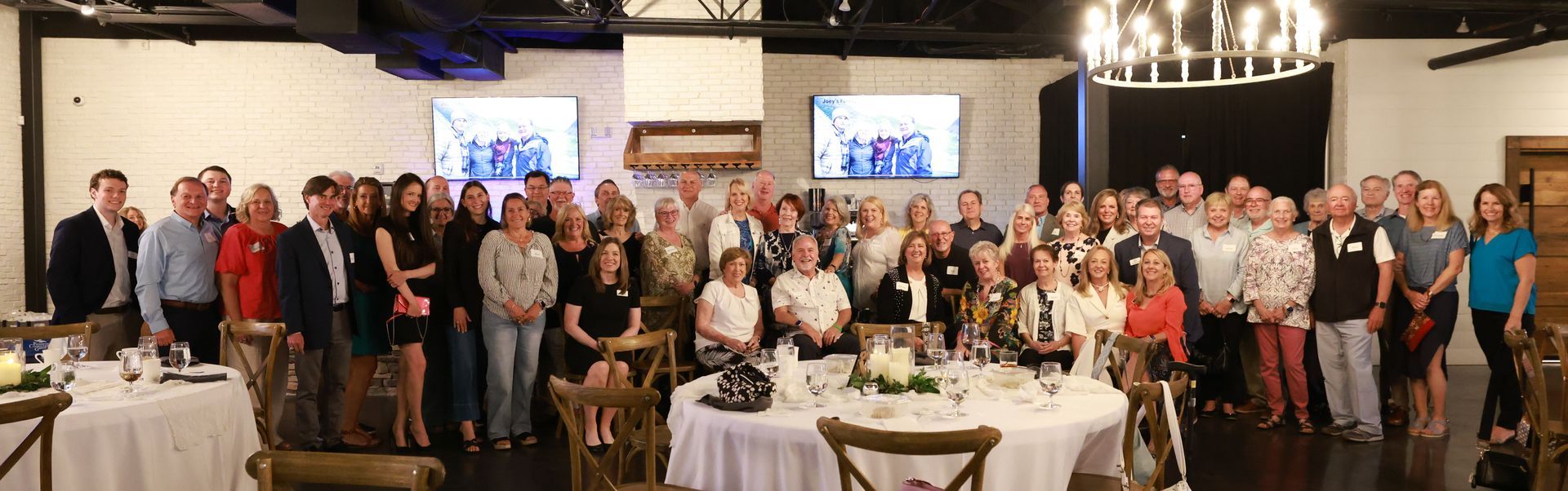 A large group of people gathered at a restaurant, standing near tables with floral centerpieces, posing for a photo.
