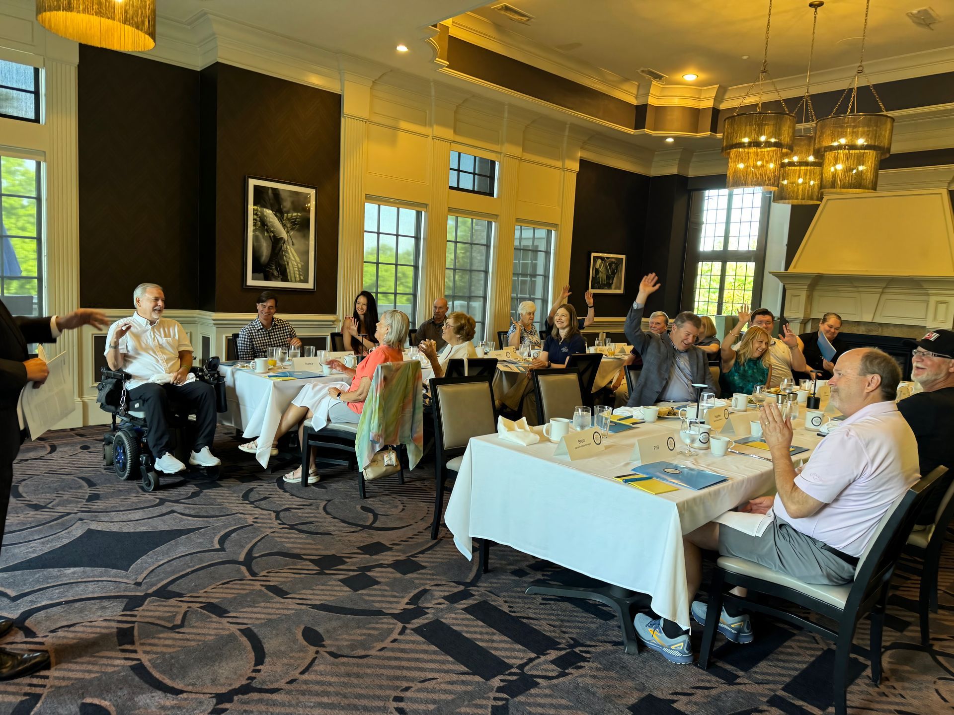 People seated at tables in a restaurant, some raising their hands. The room has large windows and ornate lighting.
