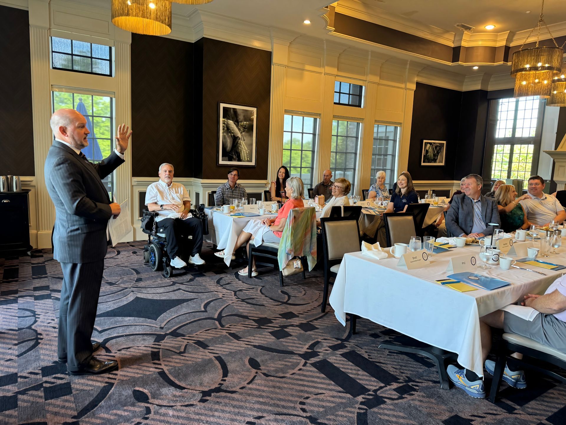 A man in a suit speaks to a seated group in a bright room. Tables are set with attendees enjoying a presentation.