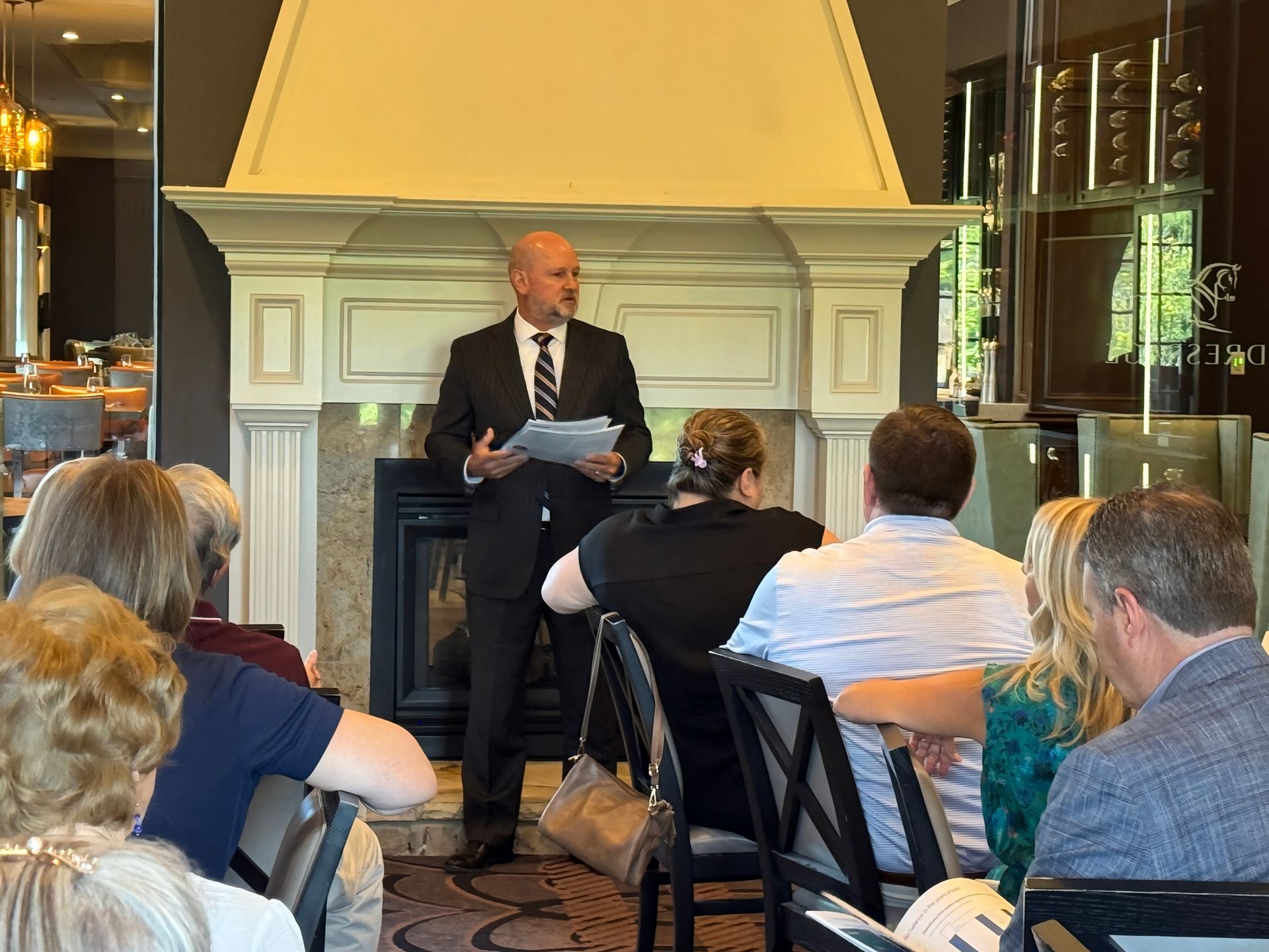 A man in a suit speaks to a seated audience in front of a fireplace. The setting appears to be an upscale restaurant.