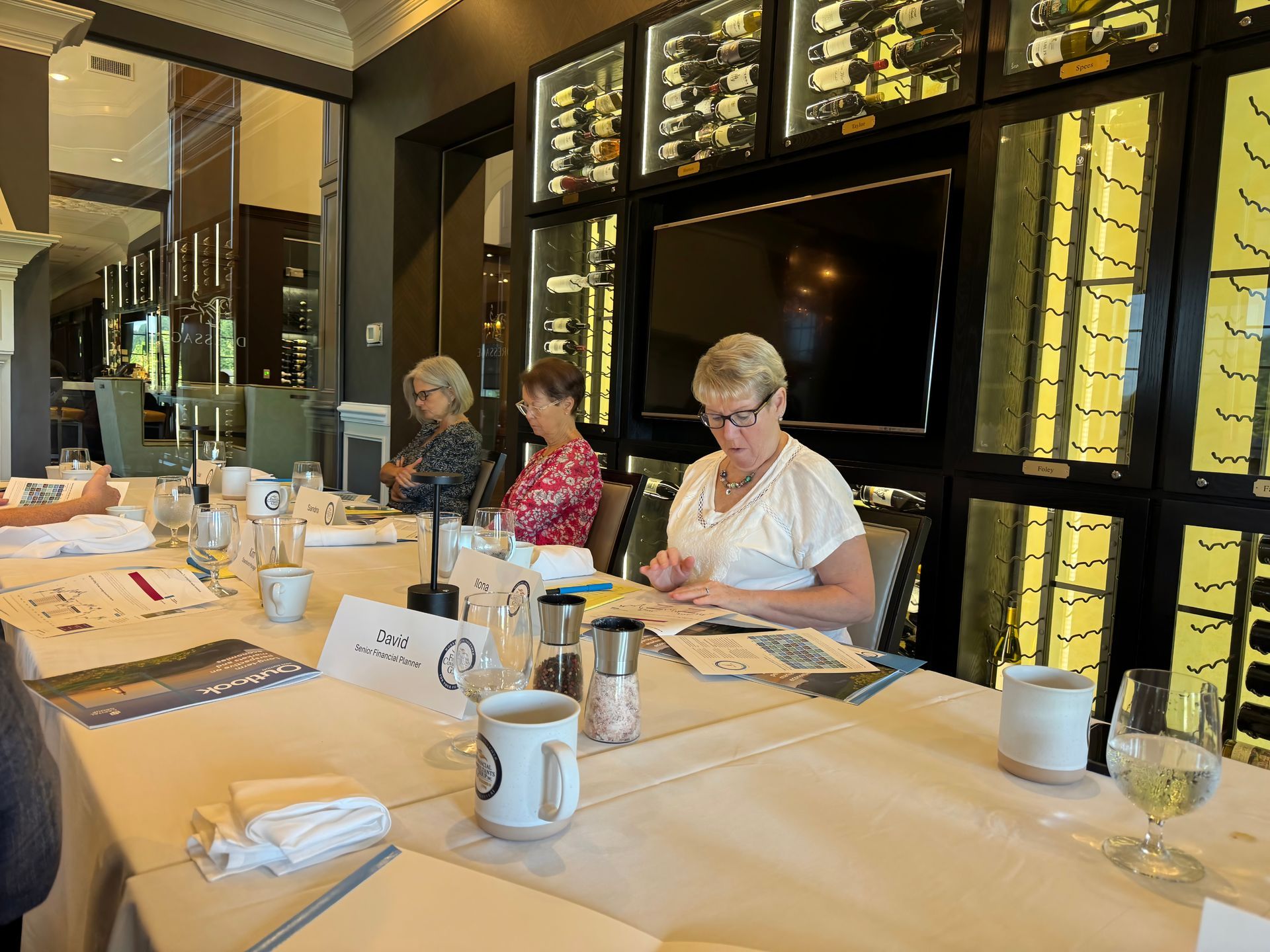 Three women seated at a long table in a restaurant, reviewing documents. Wine bottles are displayed behind them.