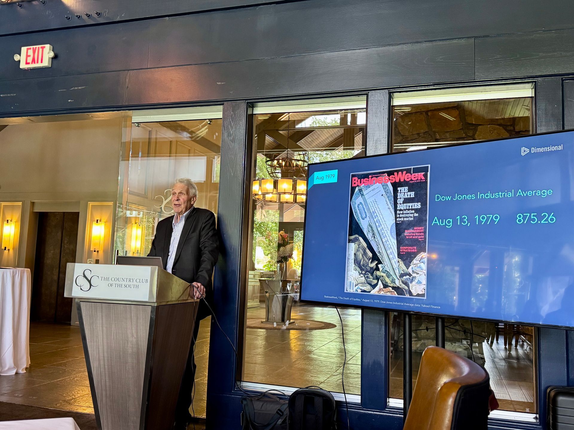 Man speaking at a podium, next to a screen displaying a poster with information. They are in a restaurant with large windows.