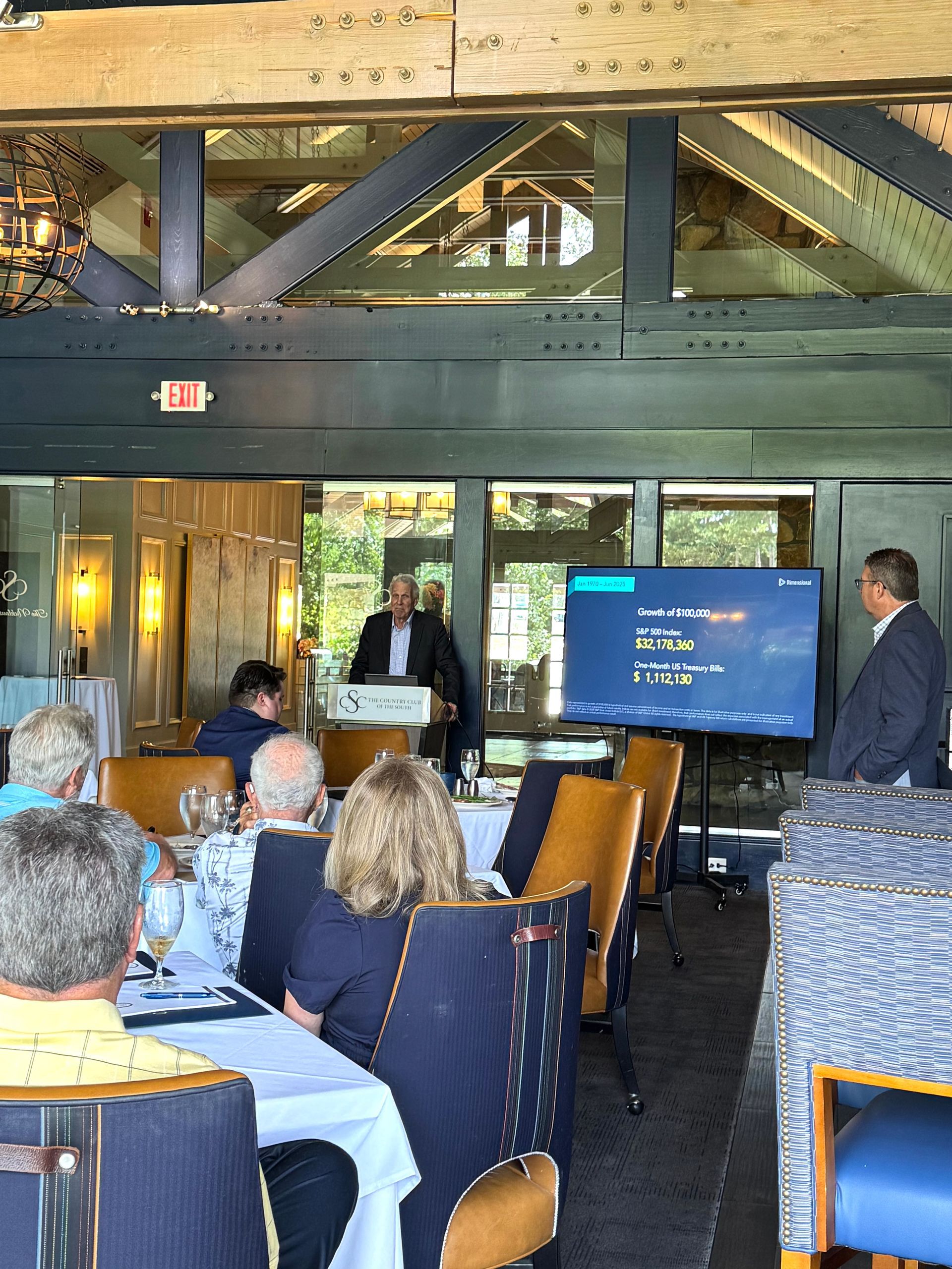 A presentation is given to a seated audience in a restaurant. Two men stand near a screen.