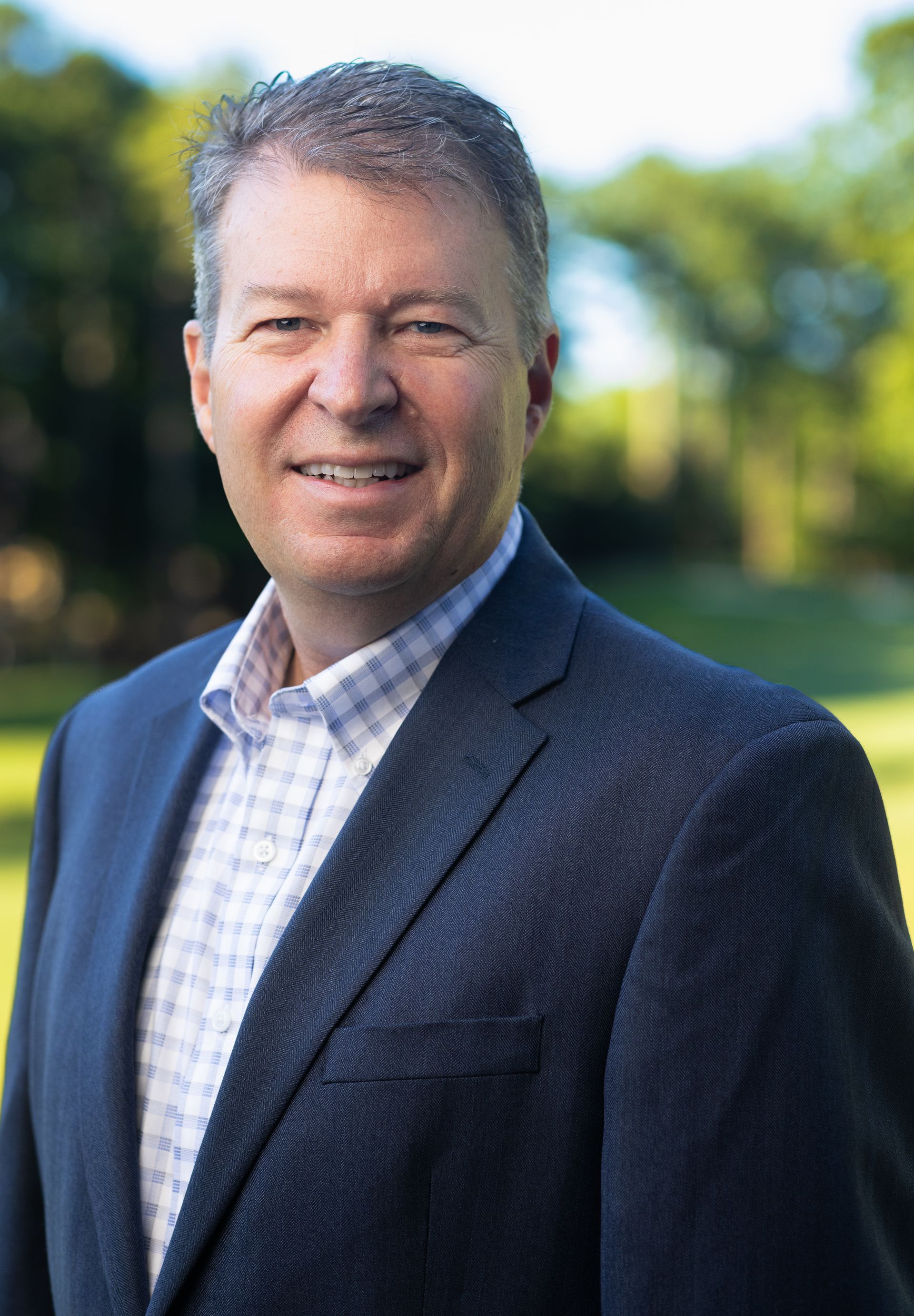 A man in a suit and white shirt is smiling in front of a brick wall.