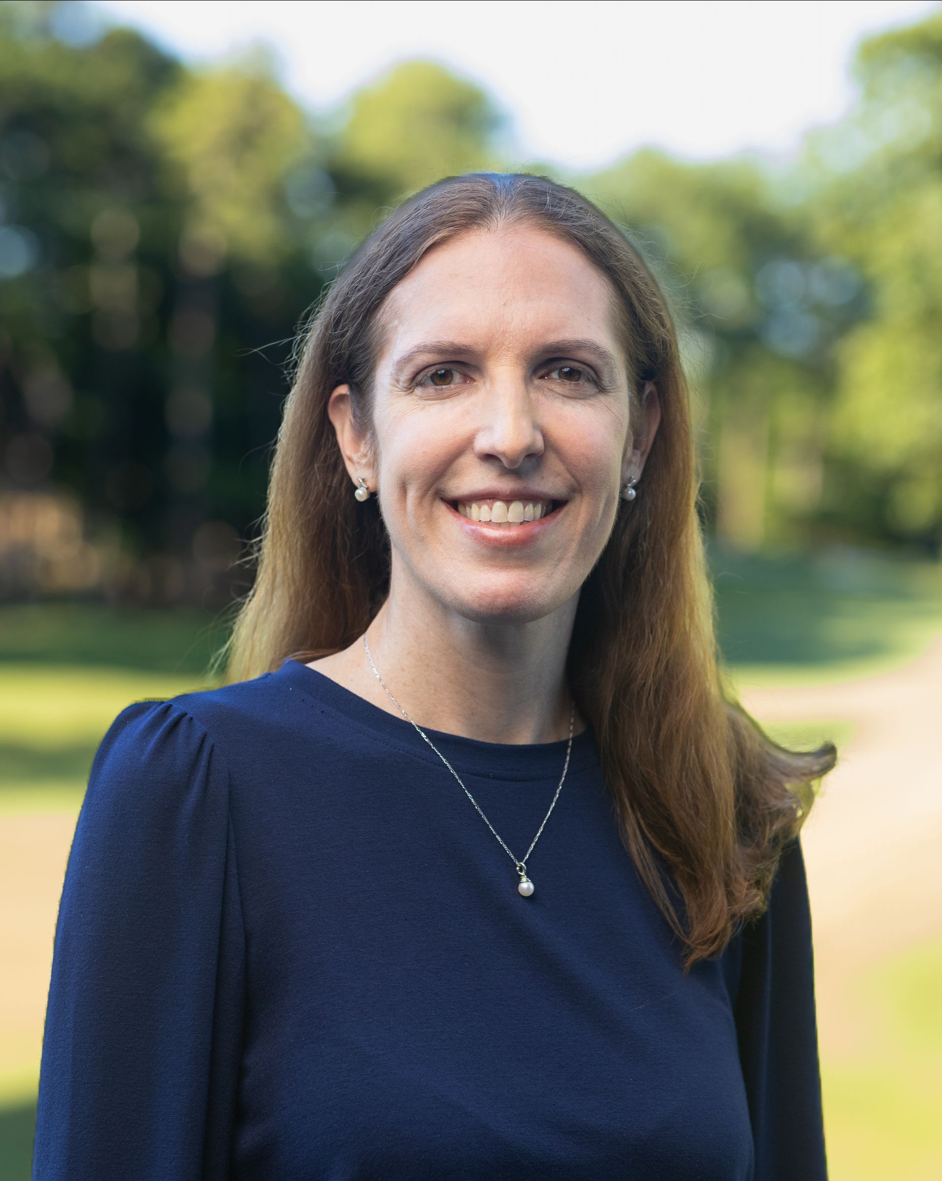 Woman with long brown hair smiles at the camera while wearing a dark blue top, a silver necklace, and small earrings. Outdoor setting with trees.