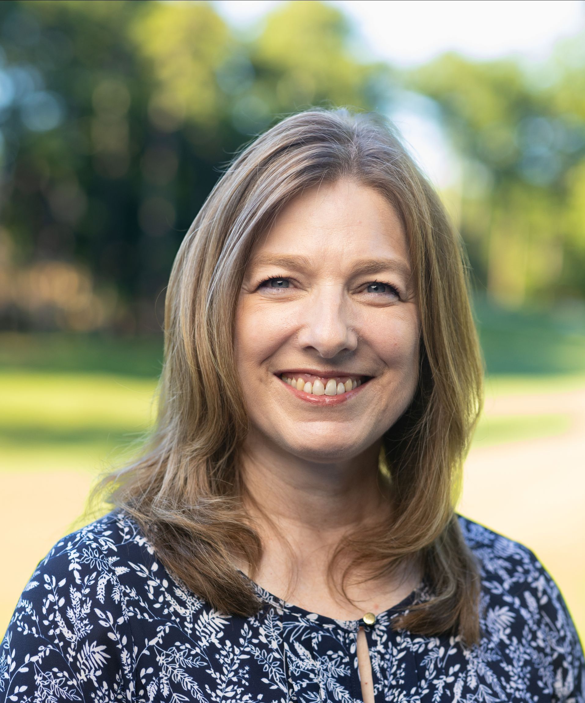Smiling woman with light brown hair, wearing a navy top, stands in a green, outdoor setting.