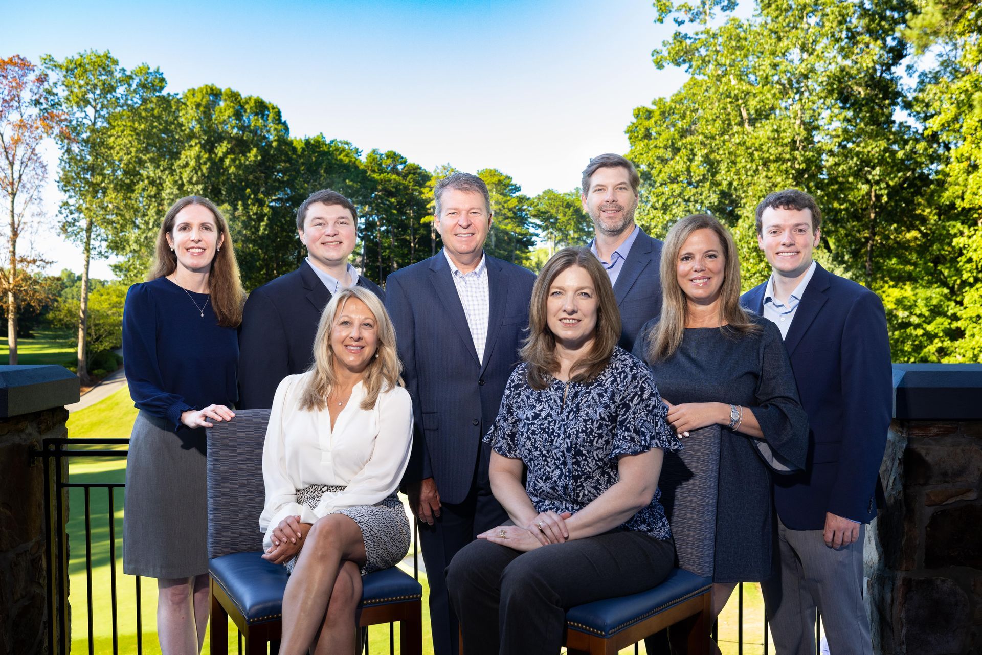 Group of nine professionals posing outdoors; some seated, some standing; trees and golf course in background.